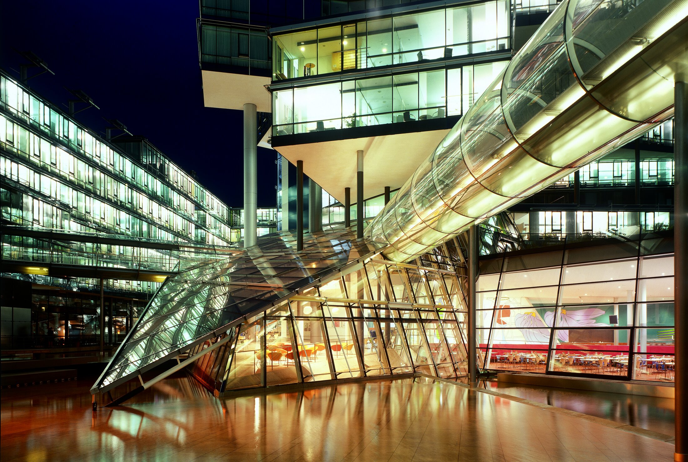 Project by Behnisch Architekturbuero, Norddeutsche Landesbank am Friedrichswall. Night view of the modern glass office complex with illuminated interiors and a tubular skybridge connecting buildings above a lit atrium.