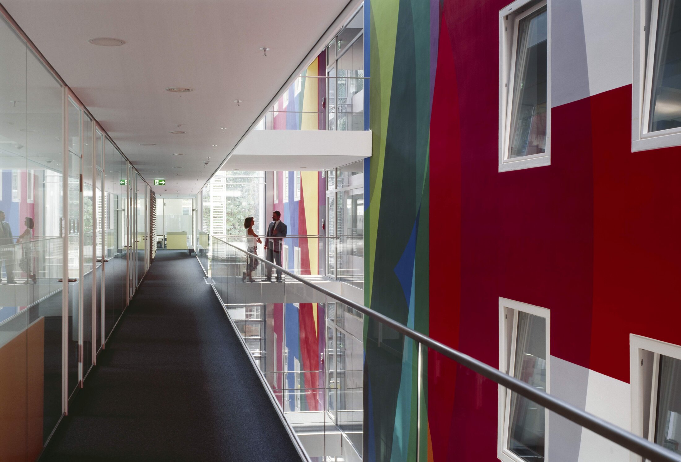 Project by Behnisch Architekturbuero, Norddeutsche Landesbank am Friedrichswall. Interior corridor with glass offices and railing, facing a colorful wall. Two people stand talking on a balcony across the atrium.