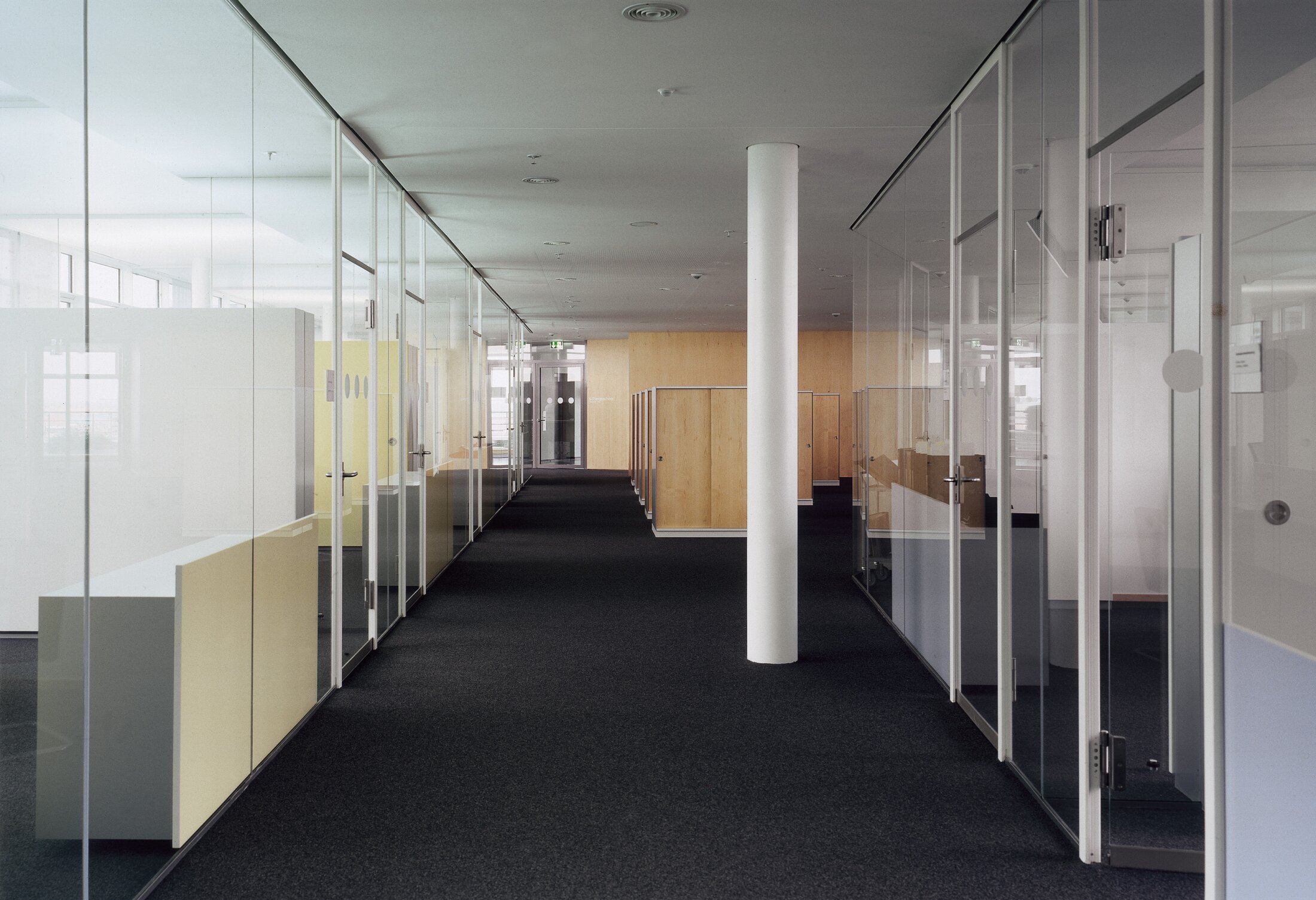 Project by Behnisch Architekturbuero, Norddeutsche Landesbank am Friedrichswall. Long office corridor with glazed rooms on both sides, a central column, dark carpeting and wooden partitions in the rear.