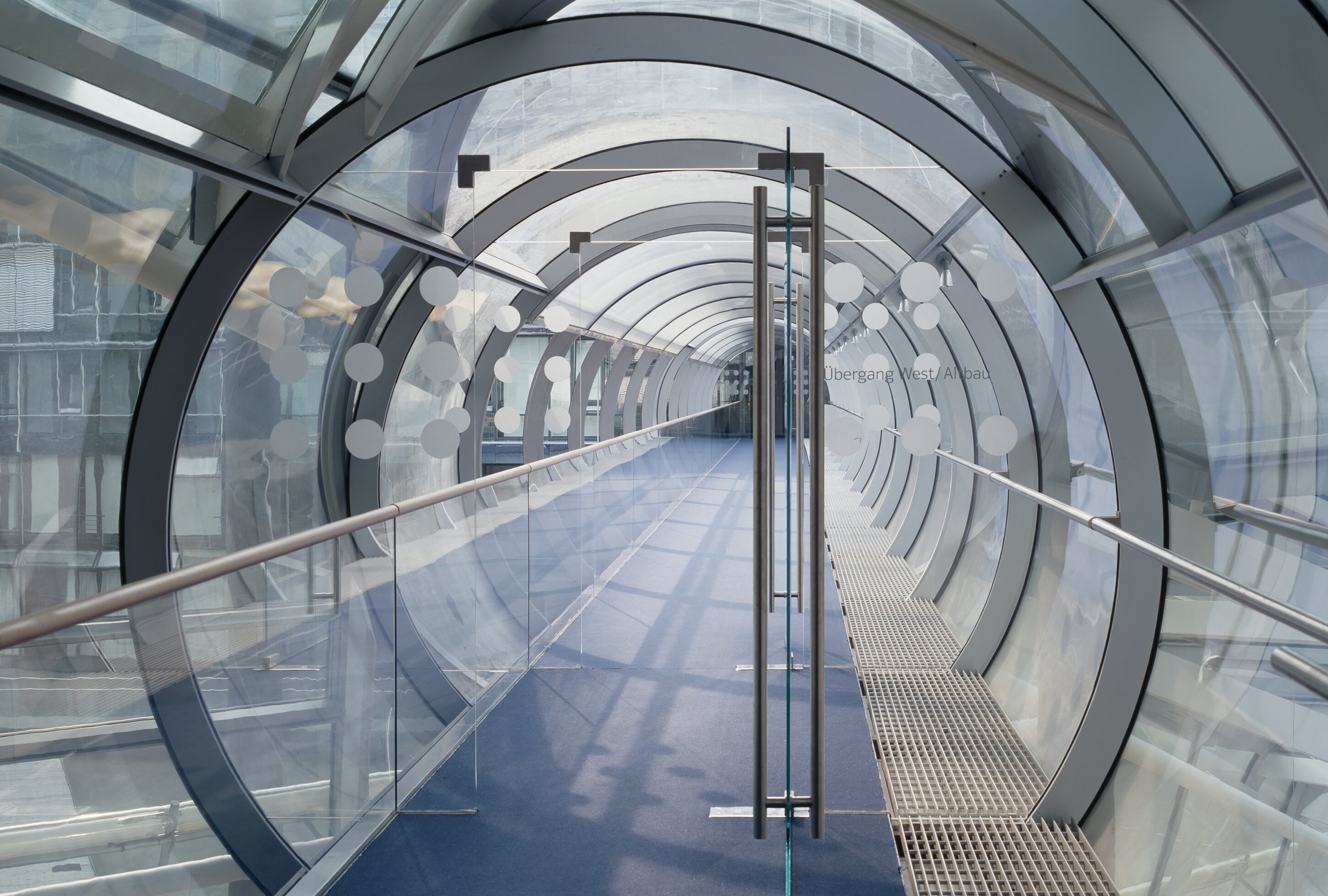 Project by Behnisch Architekturbuero, Norddeutsche Landesbank am Friedrichswall. Enclosed glass walkway with curved steel ribs, blue floor, and handrails, forming a tunnel-like corridor connecting buildings.