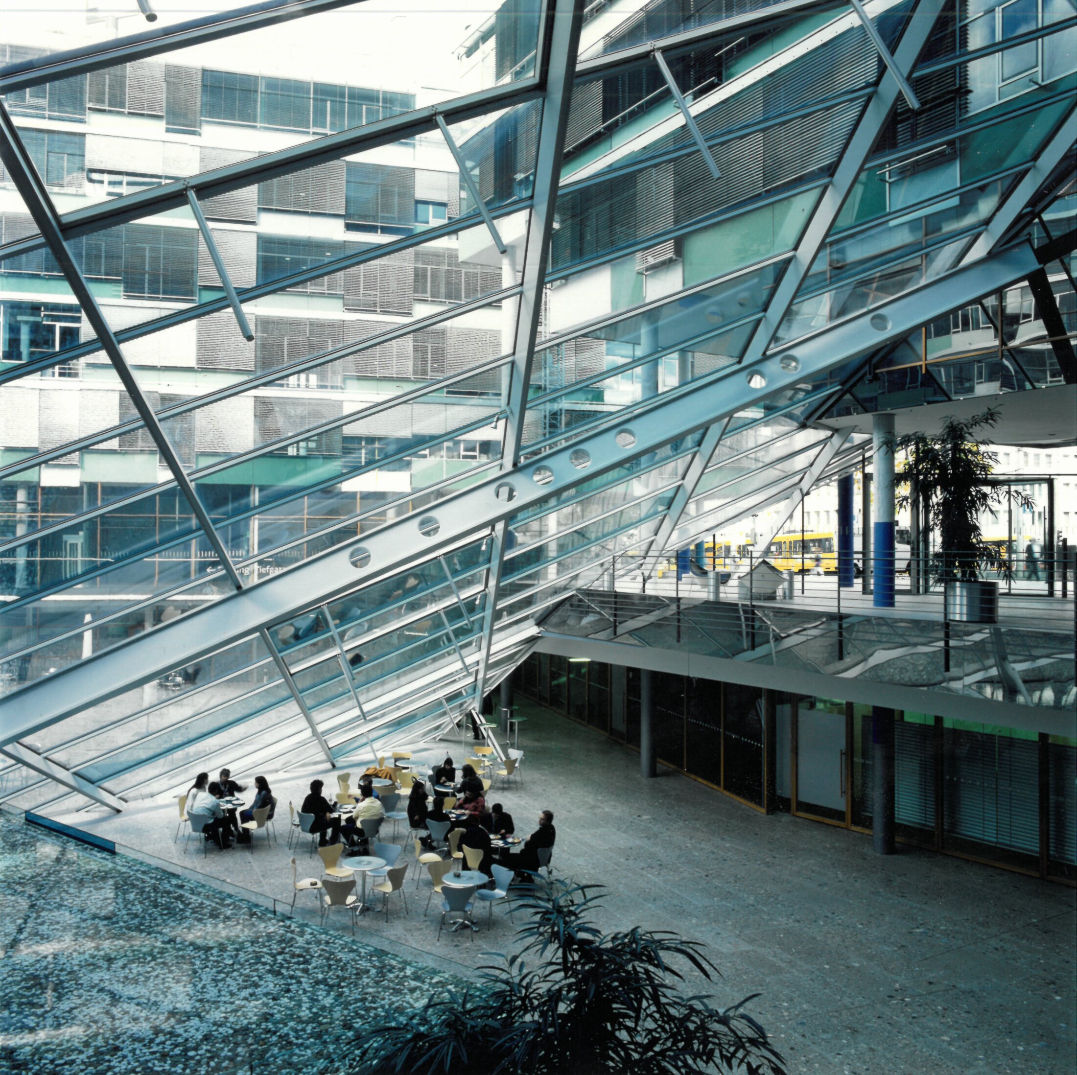 Project by Behnisch Architekturbuero, Landesgirokasse am Bollwerk. Atrium with glass roof and steel beams, greenery and people sitting at tables in a caf&eacute;-like space.