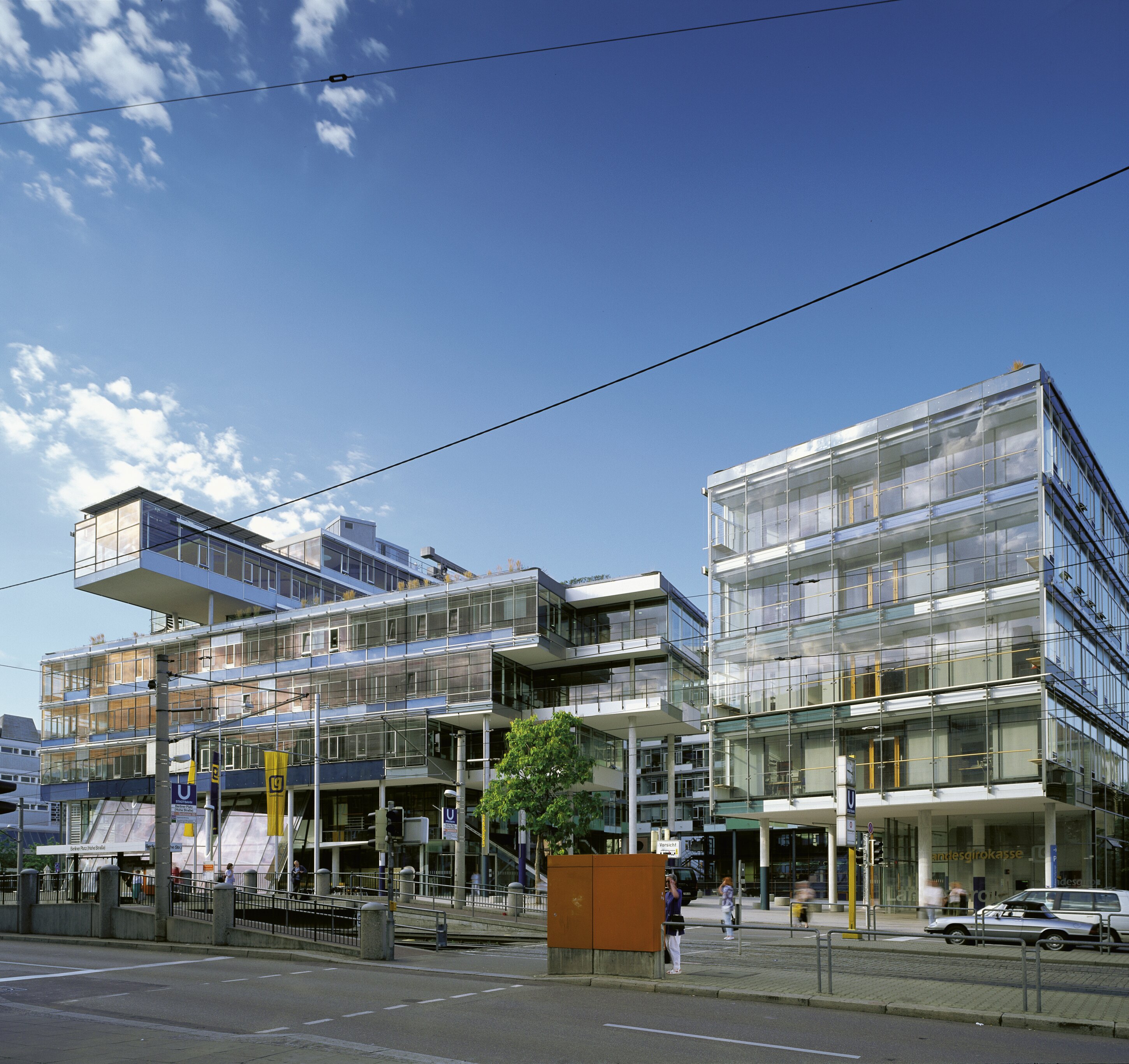 Project by Behnisch Architekturbuero, Landesgirokasse am Bollwerk. Modern glass office buildings at a city street intersection with tram wires, pedestrians, and a clear blue sky.