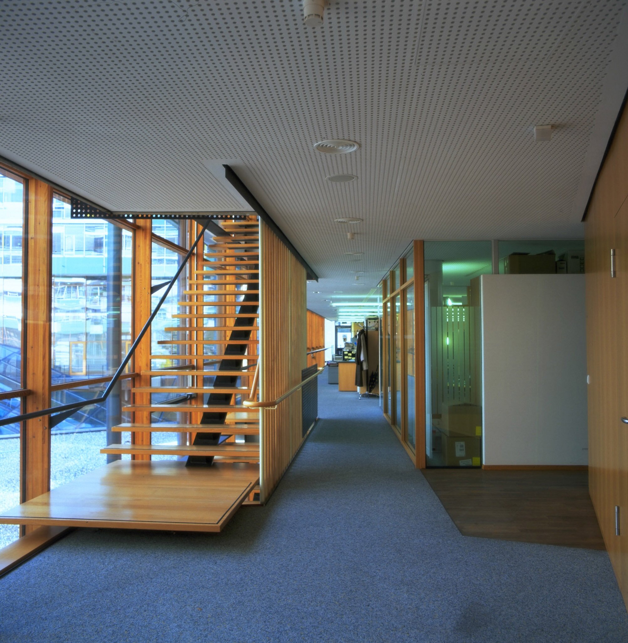 Project by Behnisch Architekturbuero, Landesgirokasse am Bollwerk. Office corridor with glass walls, wooden staircase, and carpeted floor leading past rooms and coat racks.