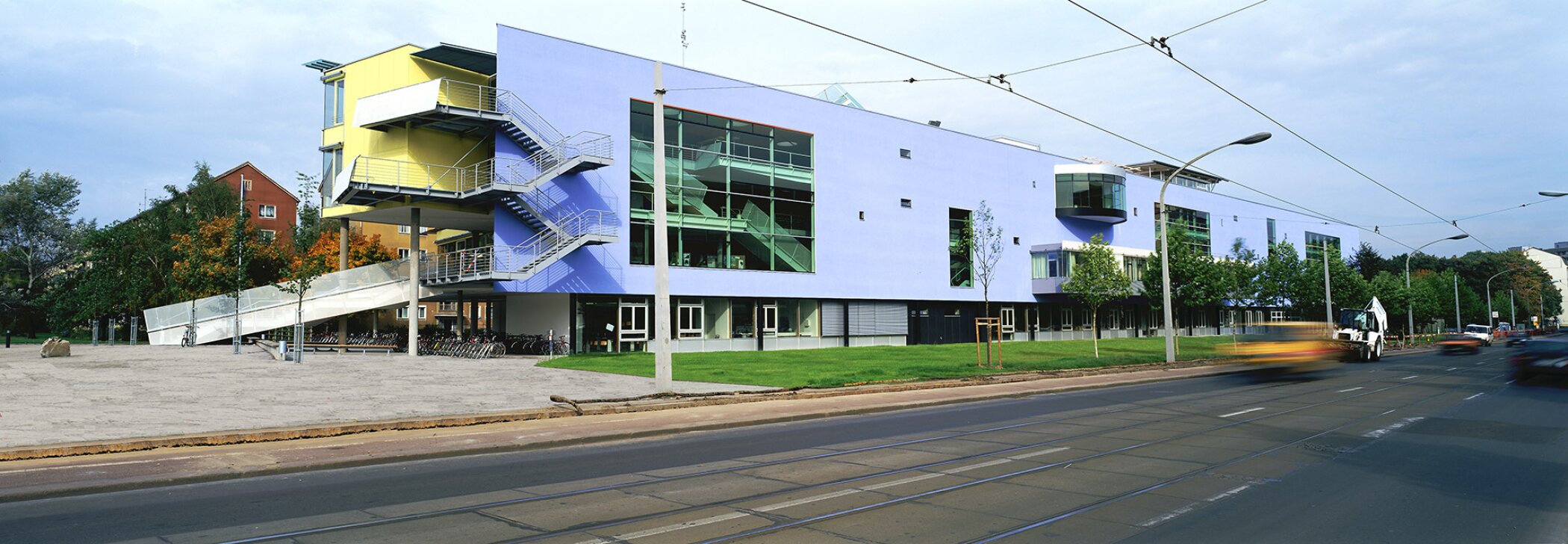 Project by Behnisch Architekturbuero, St. Benno School. Modern building with blue and yellow facade, external stairs, and large windows beside a street with tram tracks and passing cars.