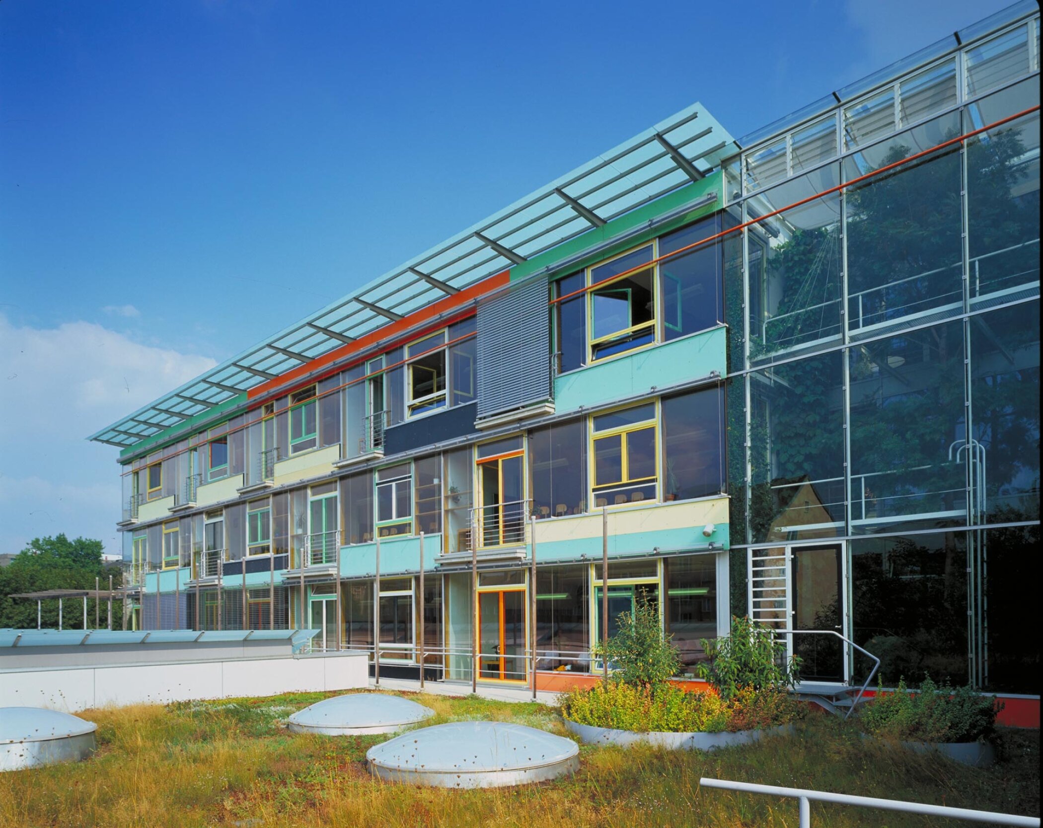 Project by Behnisch Architekturbuero, St. Benno School. Colorful modern building with glass facade, varied window frames, and rooftop canopy, seen above a planted area with round skylights.
