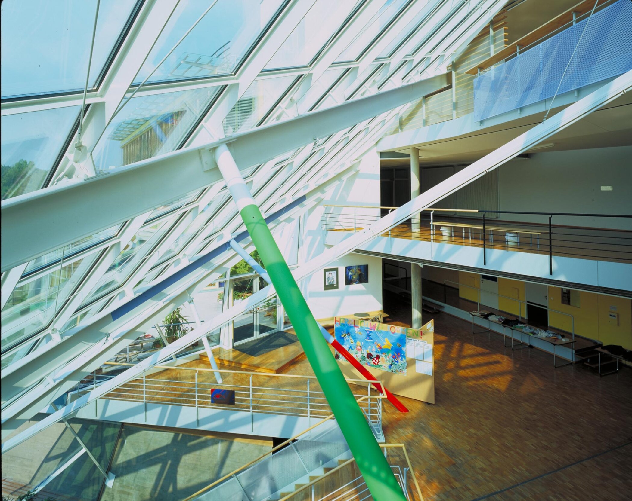 Project by Behnisch Architekturbuero, St. Benno School. Sunlit atrium with slanted glass roof, exposed beams, walkways, and a central display area on a wooden floor below.