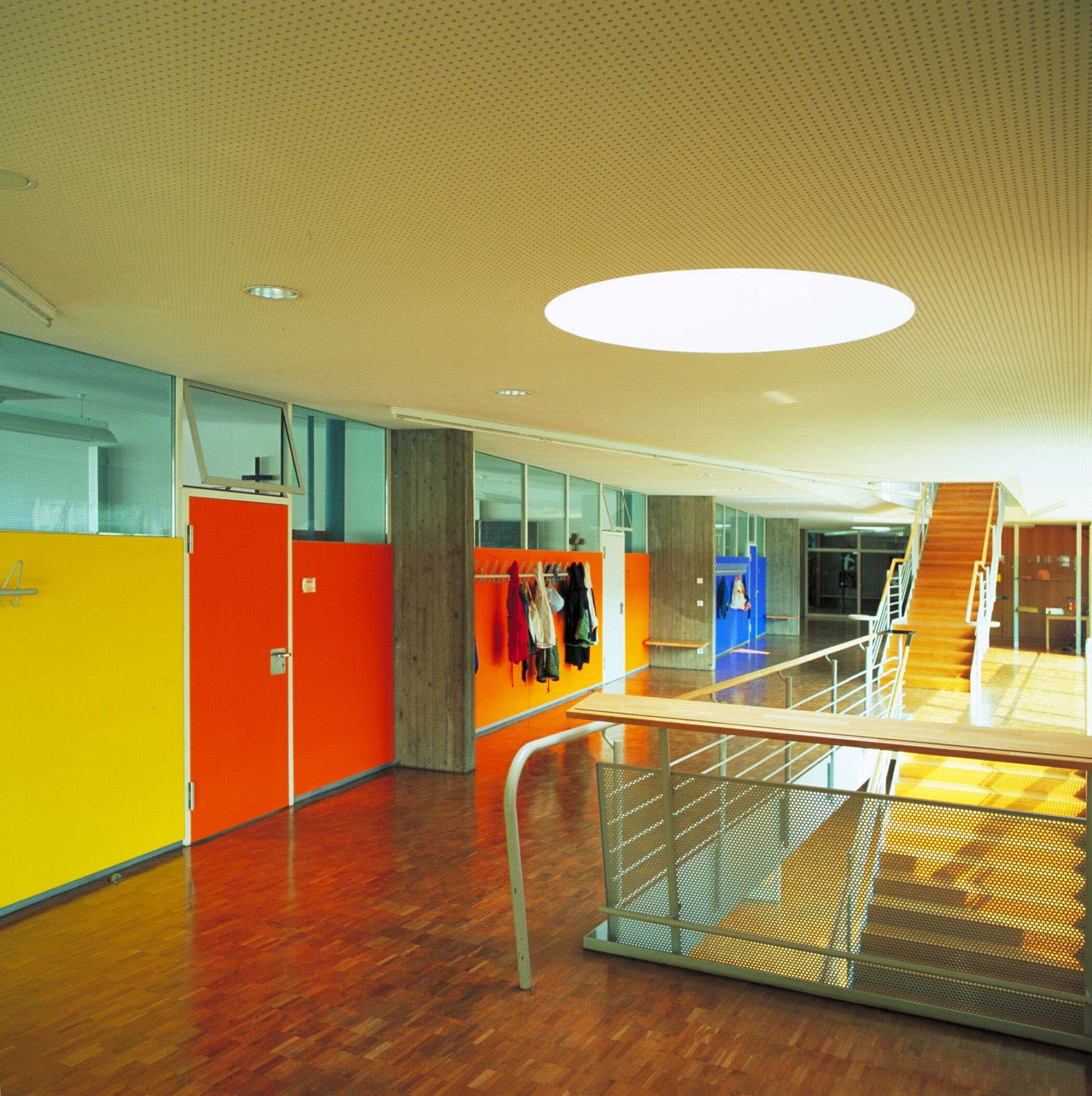 Project by Behnisch Architekturbuero, St. Benno School. Interior corridor with wood floor, colorful doors, coat racks, and a stairway, lit by a round ceiling light and skylight.