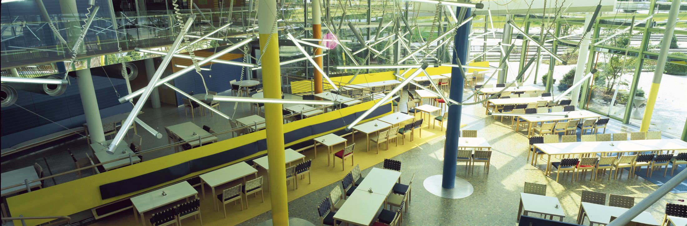 Project by Behnisch Architekturbuero, LVA &ndash; Landesversicherungsanstalt Schleswig-Holstein. Bright cafeteria with rows of tables and chairs, colorful columns, large windows, and suspended geometric light fixtures overhead.