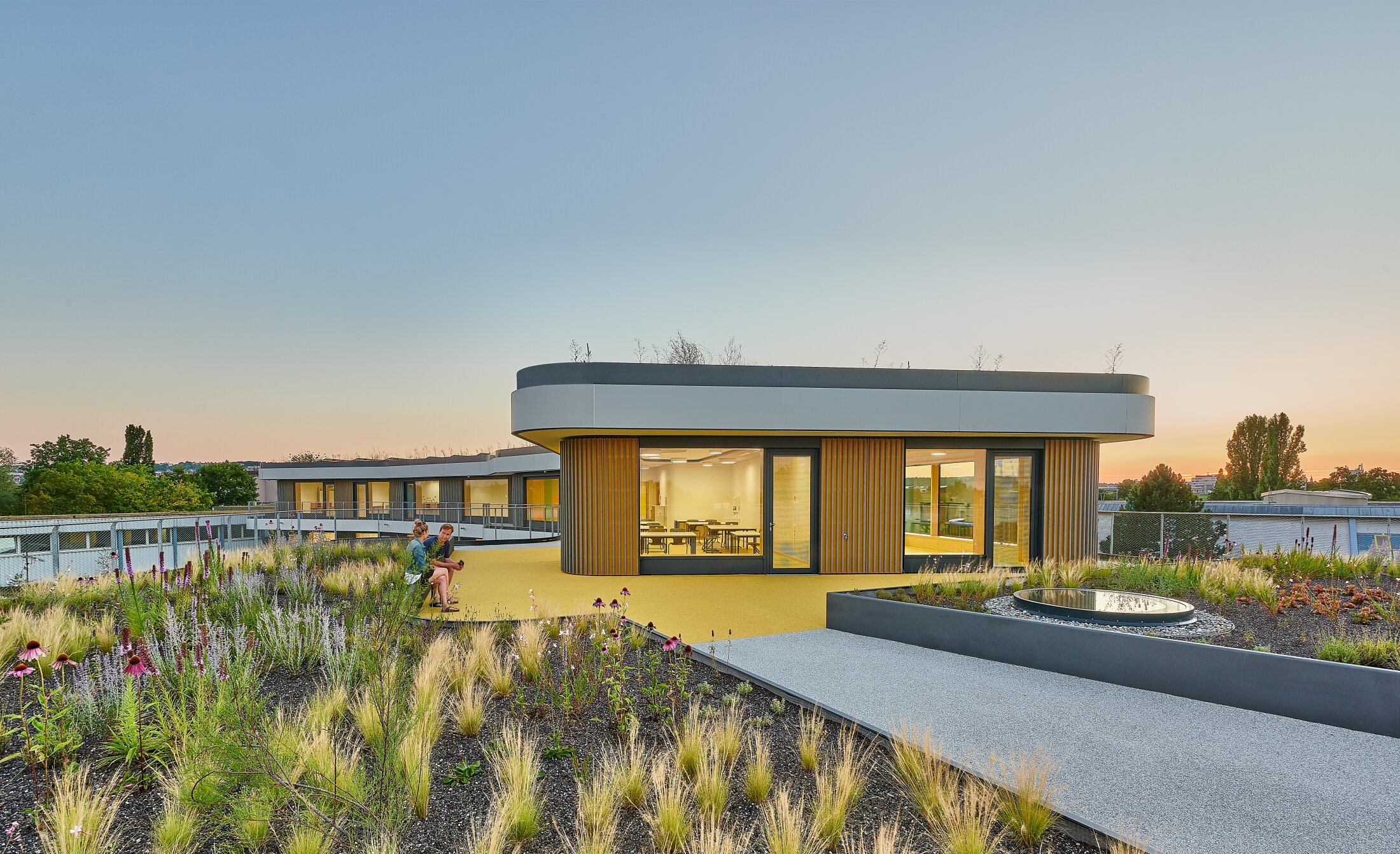 Project by Behnisch Architekturbuero, Gotthard-M&uuml;ller-School Bernhausen. A rooftop terrace with a landscaped garden and pathways; two people sit next to a modern, circular building with large windows at sunset. Part of the flooring is marked in yellow.