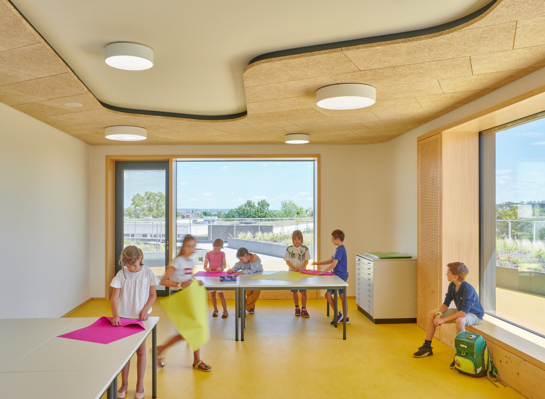 Project by Behnisch Architekturbuero, Gotthard-M&uuml;ller-School Bernhausen. Children are working at tables in a bright classroom with large windows, acoustic panels on the ceiling, and a yellow floor. The view overlooks an outdoor terrace.