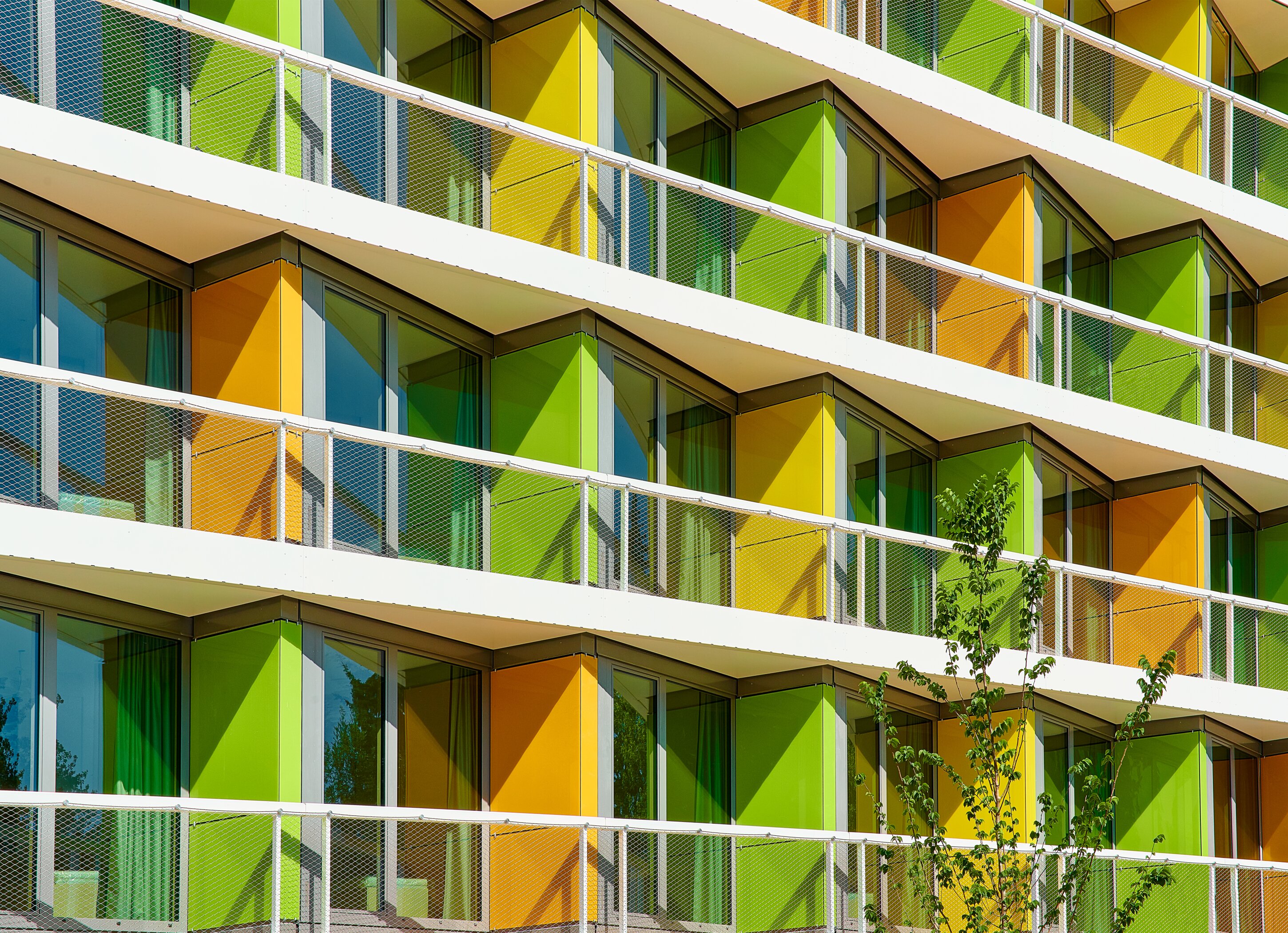 Project by Behnisch Architekturbuero, New Student Housing Complex, Nibelungenkaserne. Close-up of the building facade with repeating balconies, glass doors, and green and yellow panels, with a small tree in front.