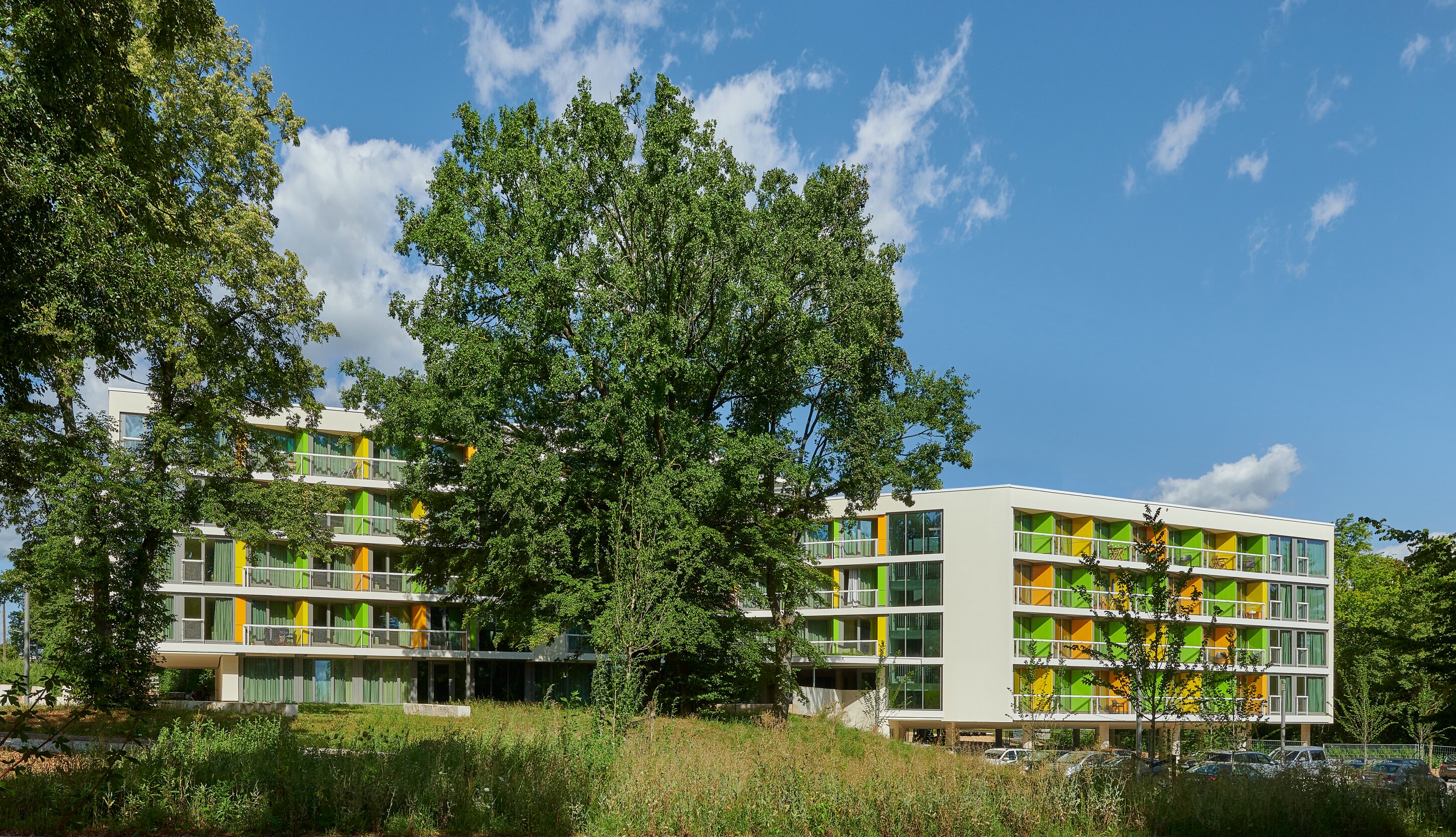 Project by Behnisch Architekturbuero, New Student Housing Complex, Nibelungenkaserne. Modern student housing with green and yellow balconies, surrounded by large trees and grass under a blue sky with scattered clouds.