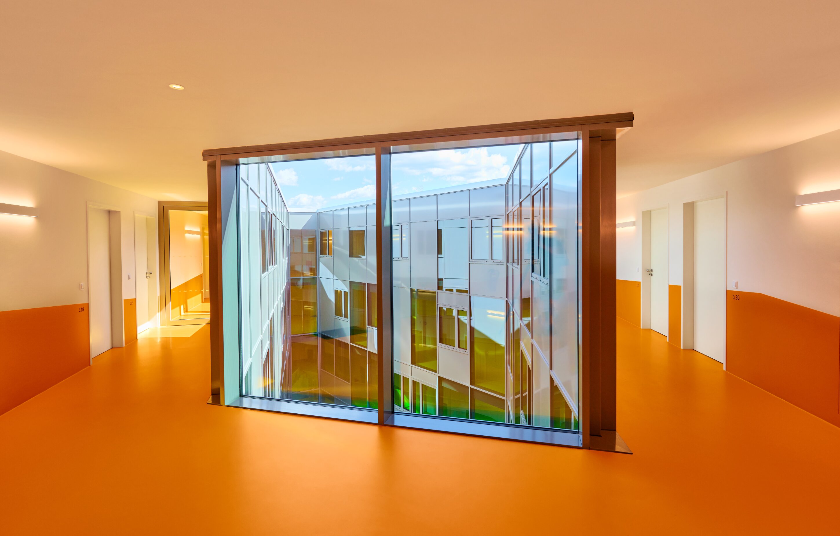 Project by Behnisch Architekturbuero, New Student Housing Complex, Nibelungenkaserne. Bright corridor with orange floor and walls, featuring a large central glass window overlooking an inner courtyard.