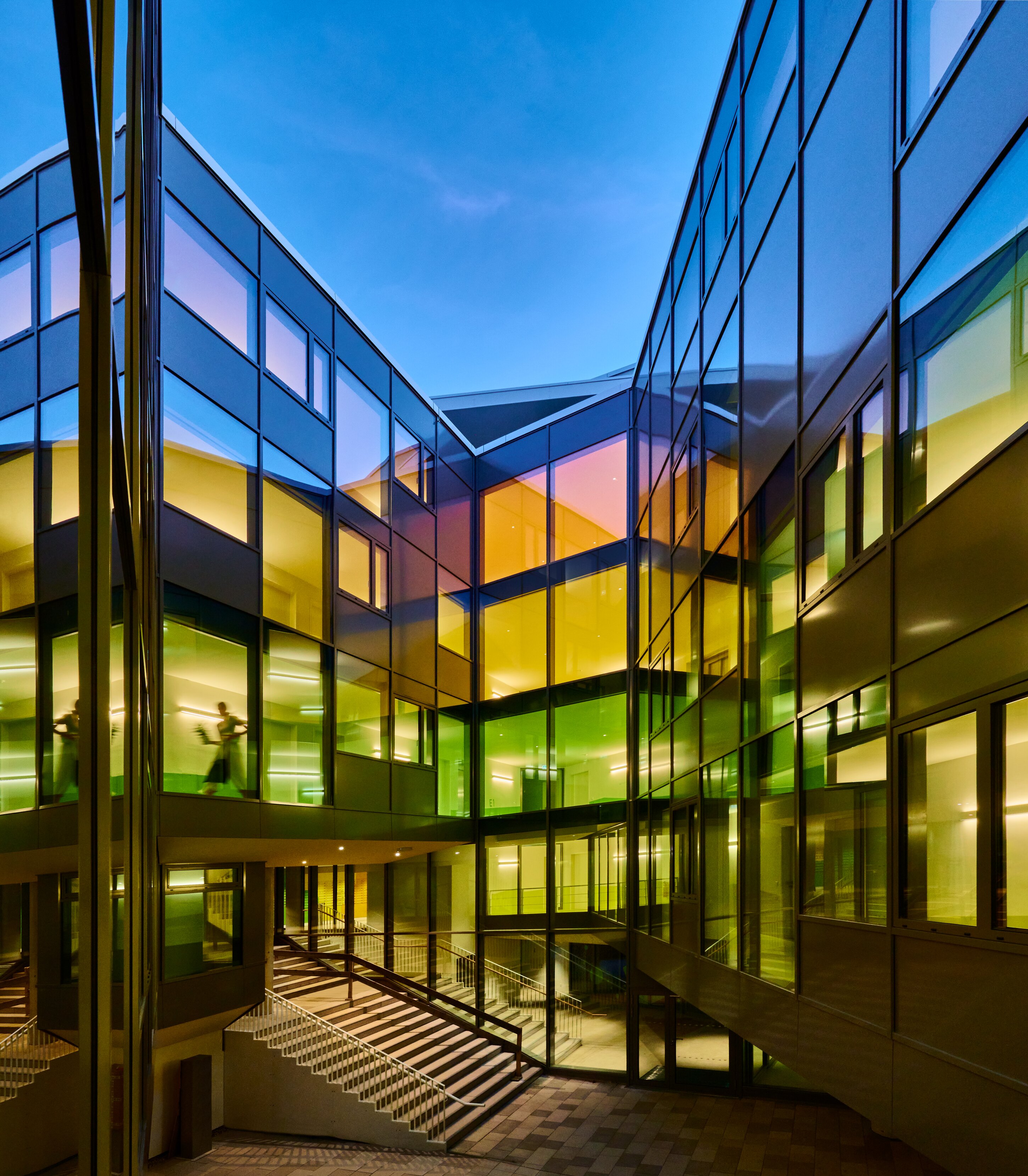 Project by Behnisch Architekturbuero, New Student Housing Complex, Nibelungenkaserne. Glass courtyard between modern buildings at dusk, with colorful lit windows and stairs leading to upper levels.
