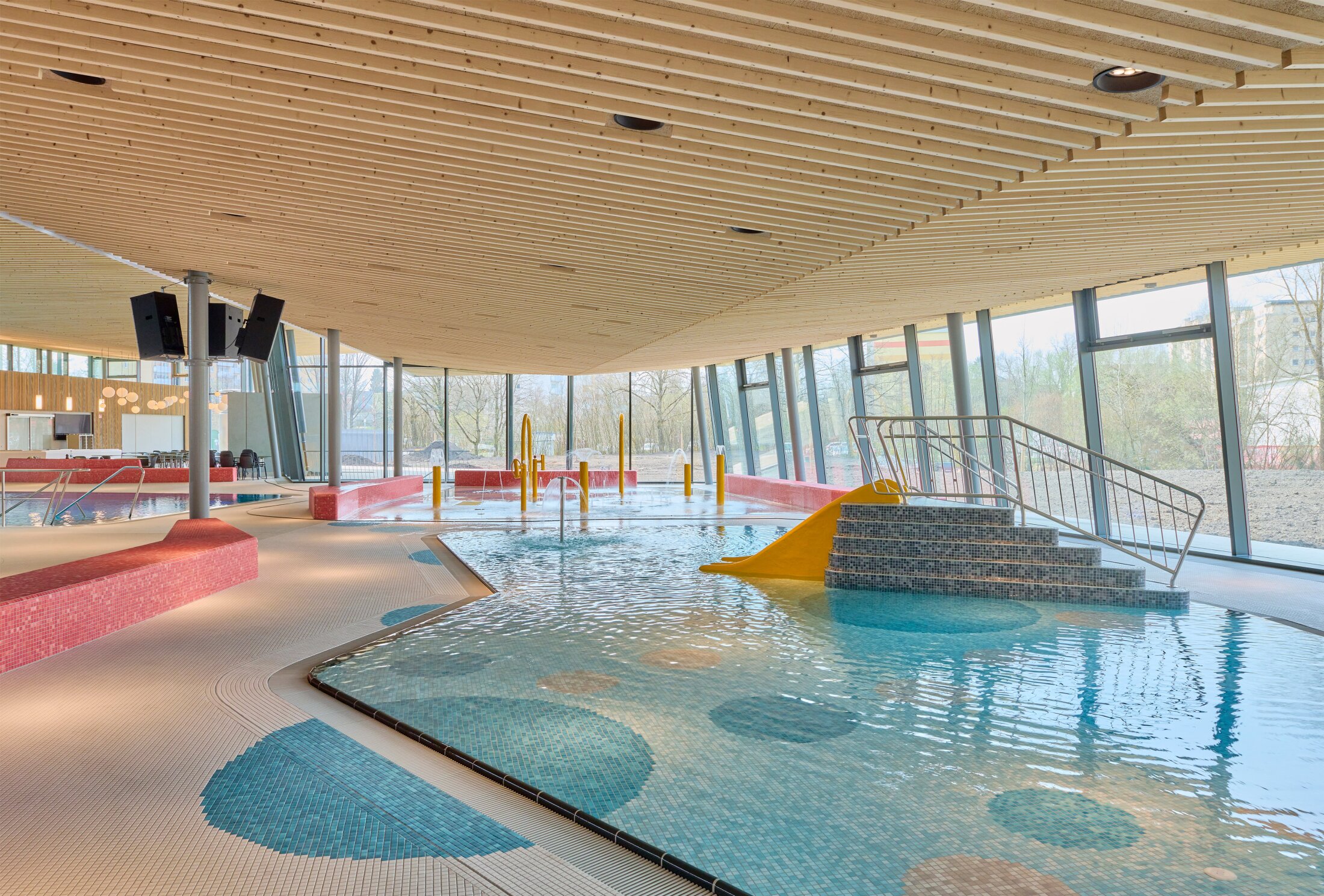 Project by Behnisch Architekturbuero, Schwaketenbad. Indoor leisure pool with shallow water, a small slide, play equipment, and large windows beneath a wooden slatted ceiling. Accents of blue, red, and yellow provide color accents.
