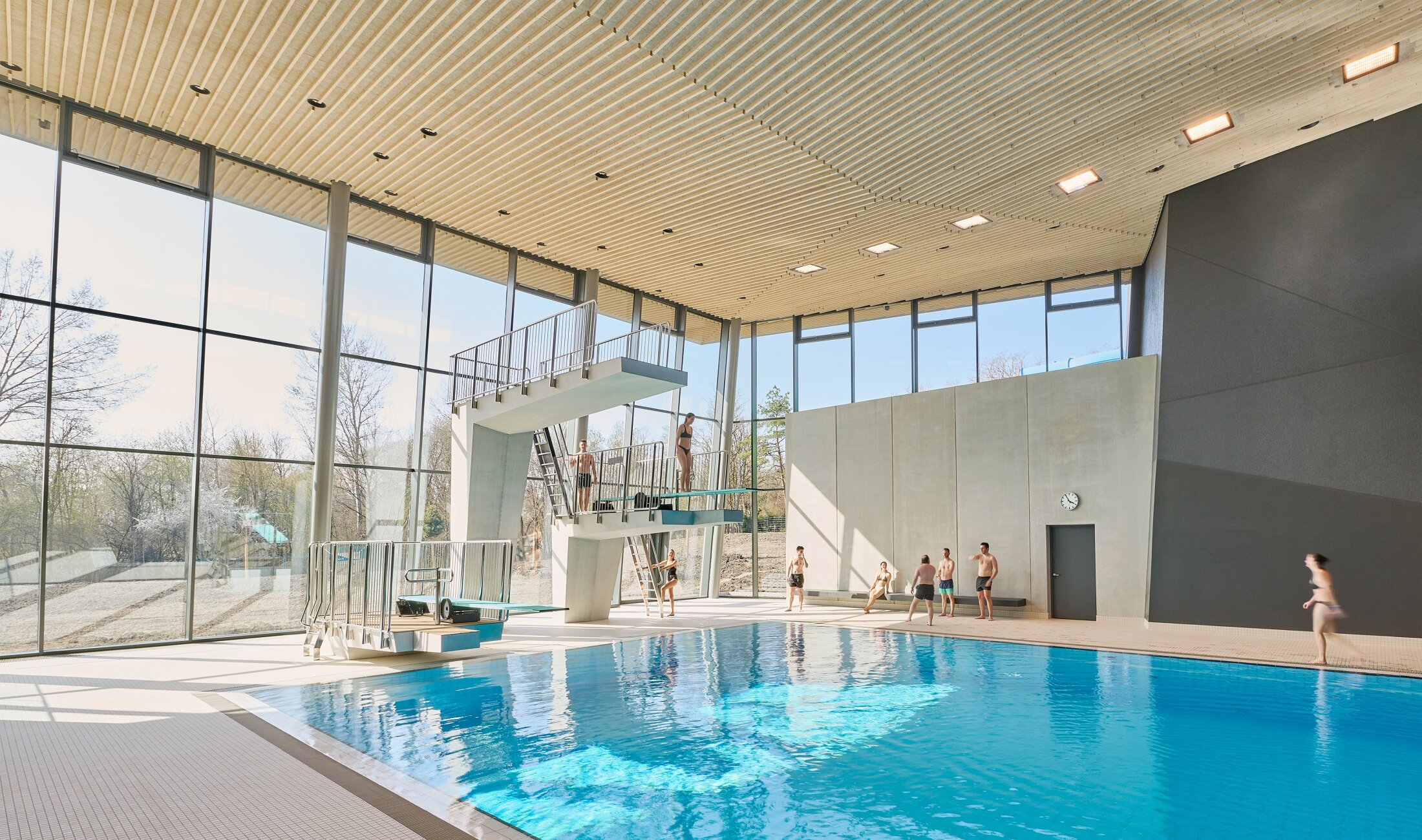 Project by Behnisch Architekturbuero, Schwaketenbad. Indoor pool with diving platforms, people standing and preparing to jump, large windows, and a high wood slatted ceiling.