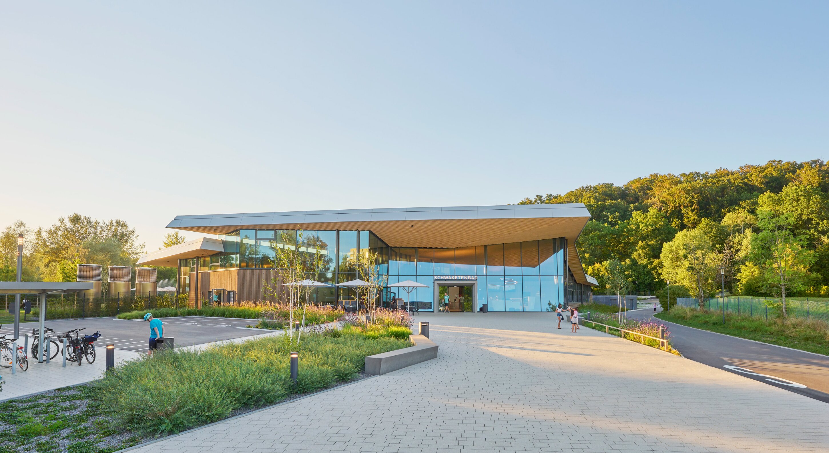 Project by Behnisch Architekturbuero, Schwaketenbad. A modern building with a glass facade, sloping roof, green plaza, bicycles and people outside, surrounded by trees and a hill. The canopy emphasizes the inviting entrance area.