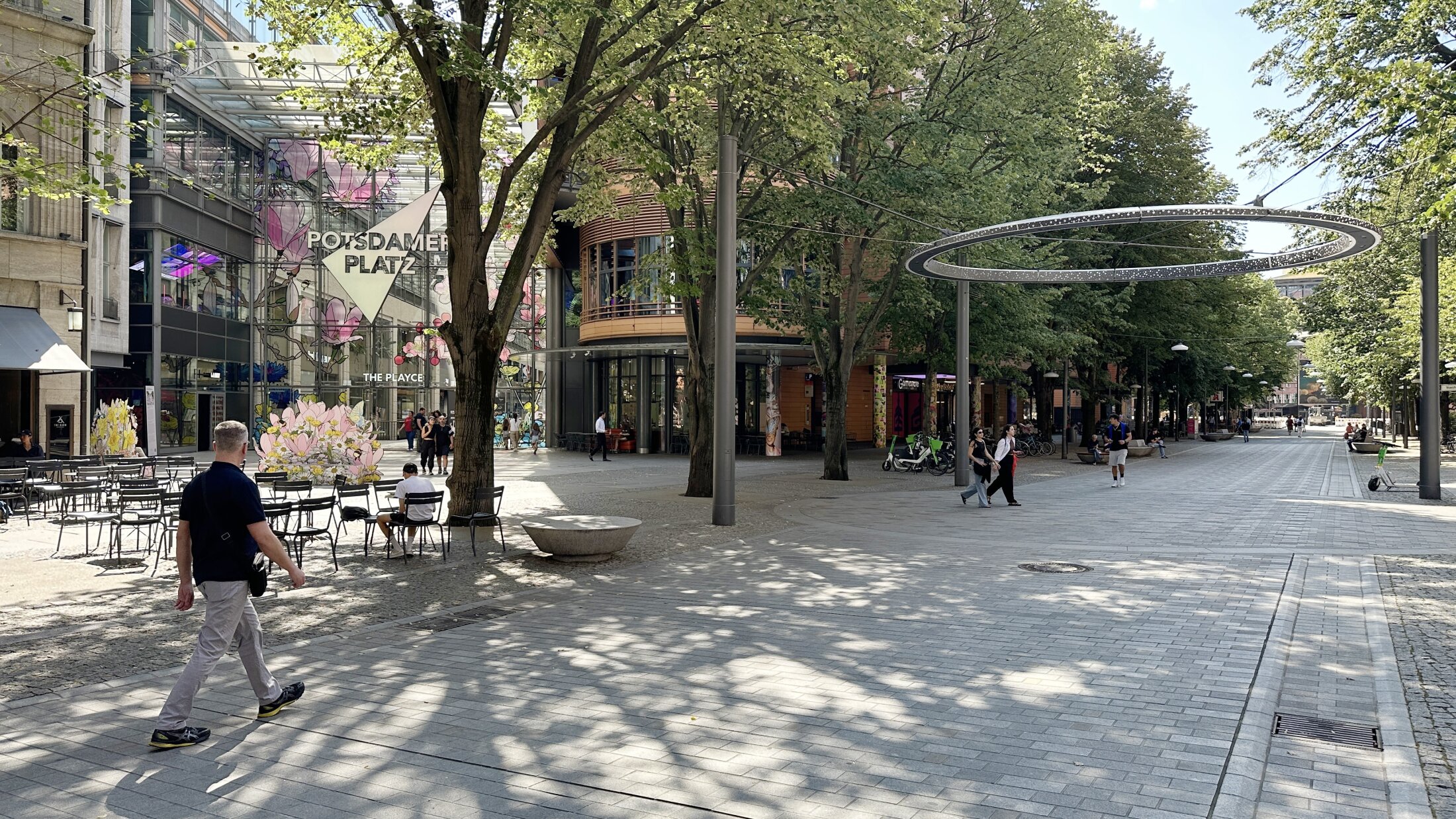 Project by Behnisch Architekturbuero, Potsdamer Platz Quartier. The tree-lined pedestrian zone at Potsdamer Platz with cafes, seating areas and pedestrians. The glass entrance can be seen in the background.