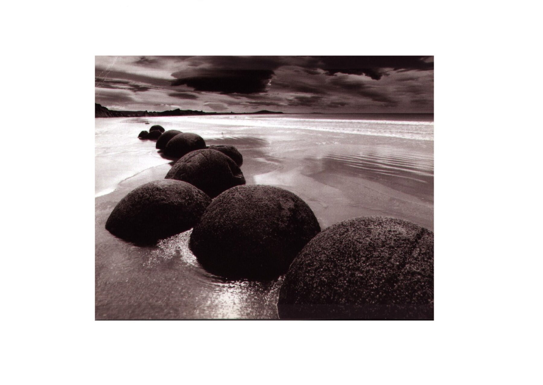 Project by Behnisch Architekturbuero, Ozeaneum. Row of large round boulders on a wet sandy beach, with shallow water, distant shoreline, and cloudy sky.