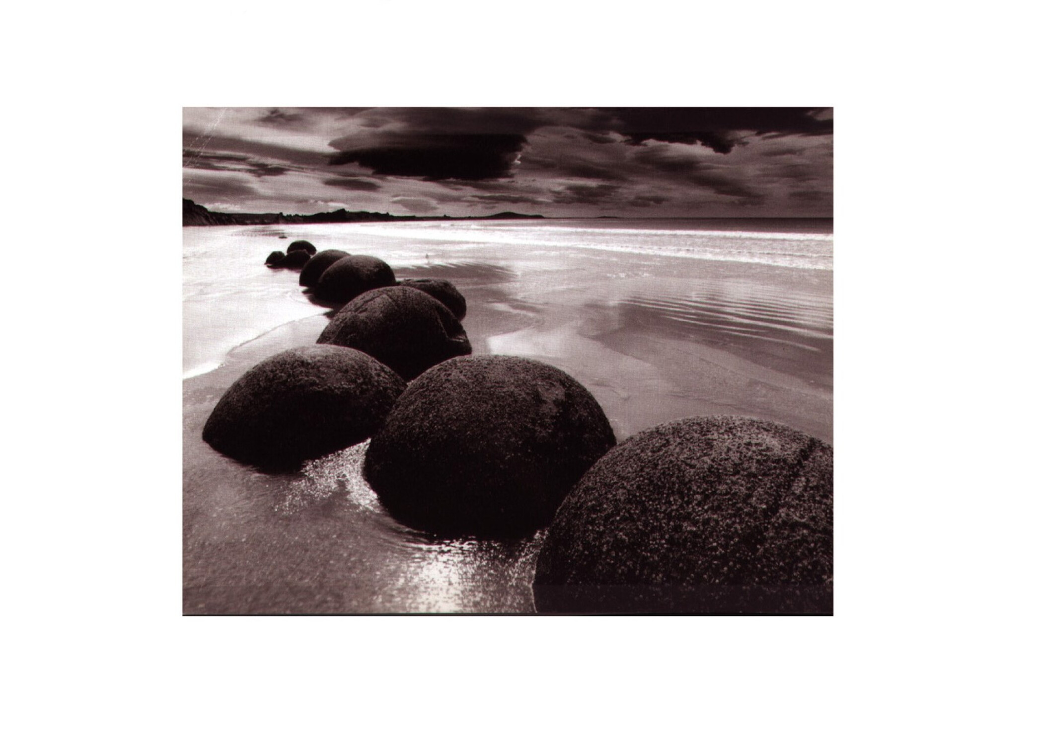 Project by Behnisch Architekturbuero, Ozeaneum. Row of large round boulders on a wet sandy beach, with shallow water, distant shoreline, and cloudy sky.