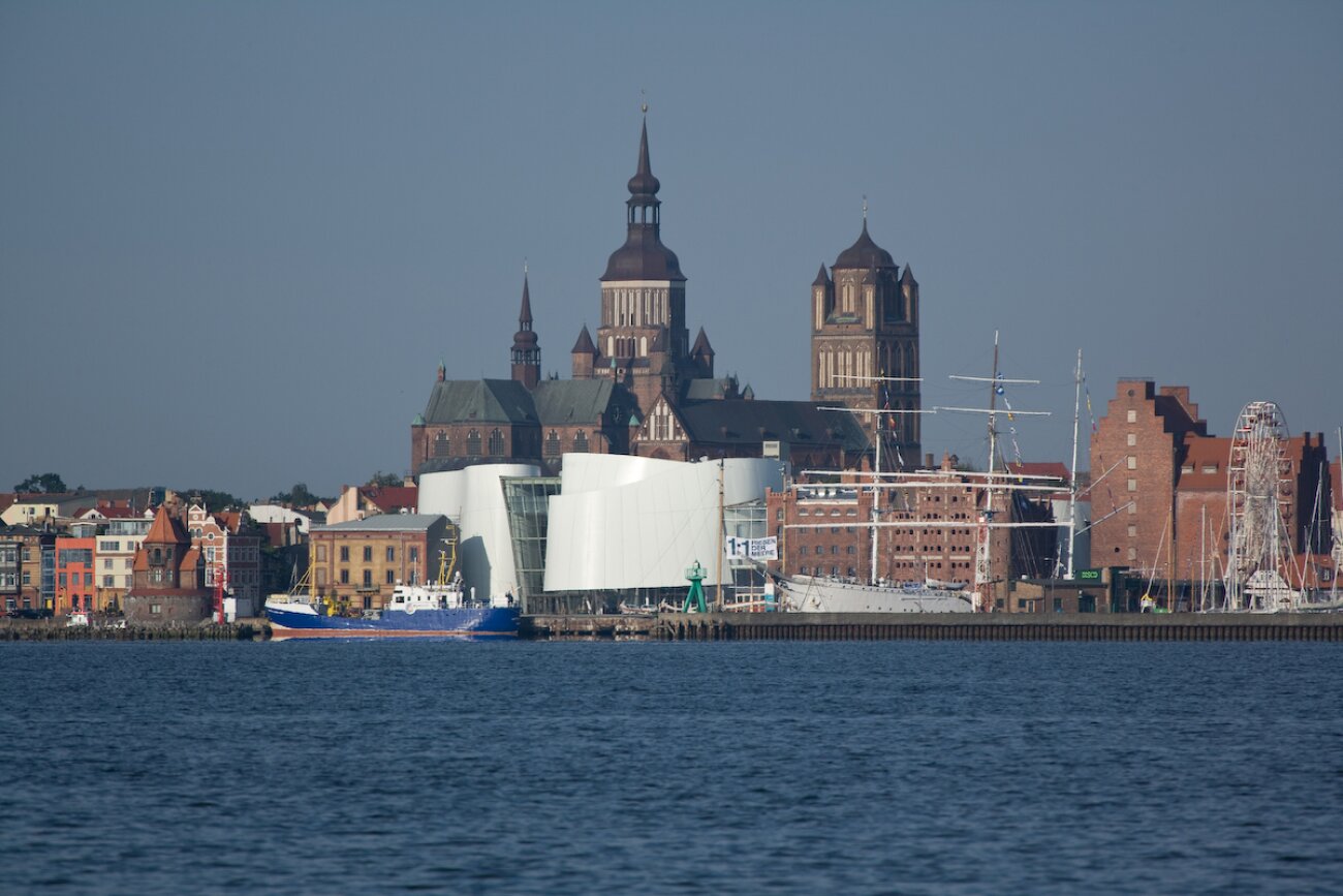 Project by Behnisch Architekturbuero, Ozeanum. Stralsund's historic waterfront promenade, with its historic church towers, anchored ships, and harbor facilities, can be seen from the opposite bank. The modern white Ozeanum building is located in the center of the image.