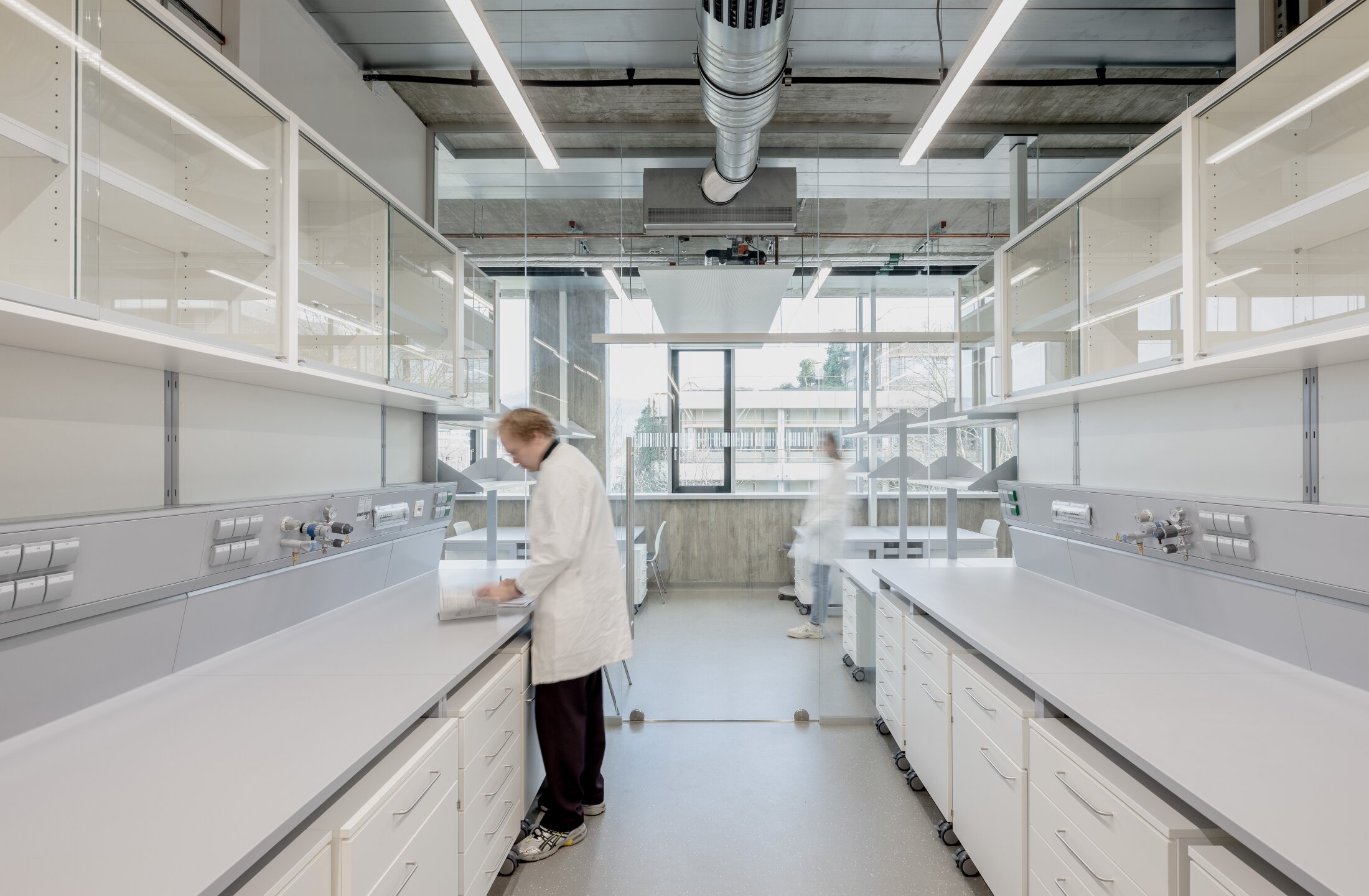 Project by Behnisch Architekturbuero, NCT New Building and Expansion. Laboratory interior with long benches and glass cabinets; two people in lab coats working at stations.