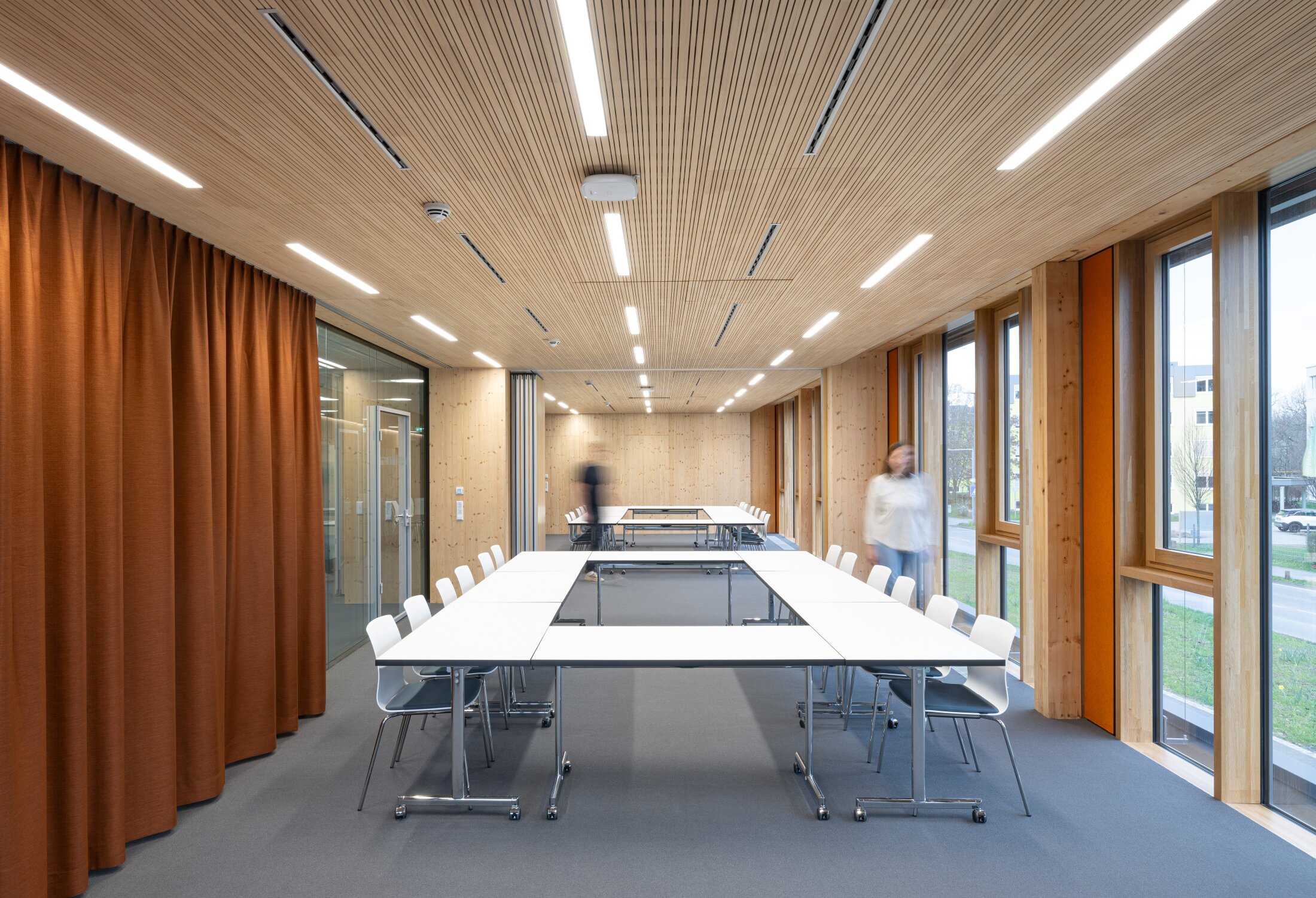 Project by Behnisch Architekturbuero, NCT New Building and Expansion. A meeting room with tables, chairs, a wooden ceiling, large windows, and people moving through the room. Color accents are provided by the orange/ochre curtain and the wall covering.