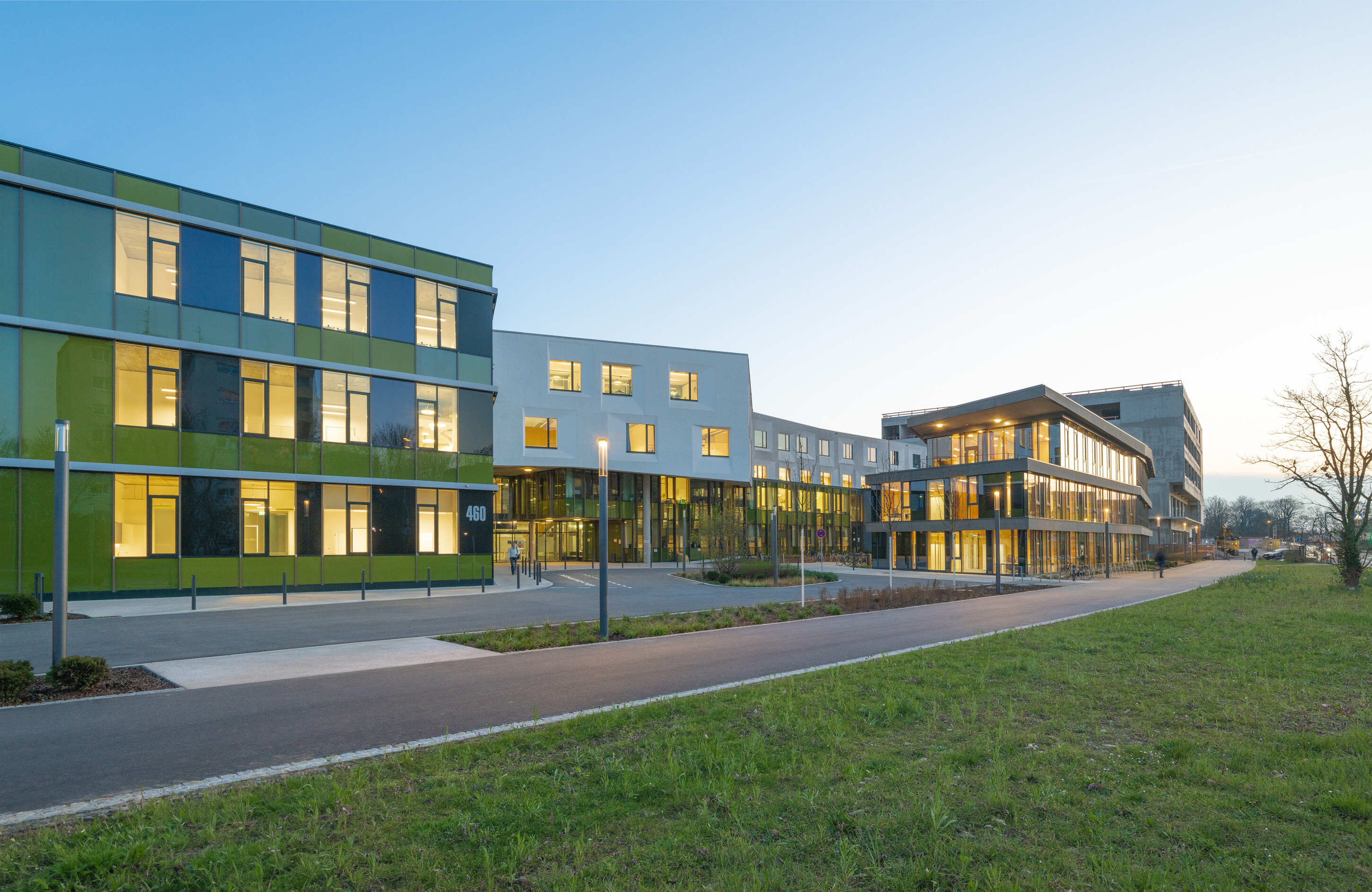 Project by Behnisch Architekturbuero, NCT New Building and Expansion. Modern campus buildings with glass facades lit at dusk, set along a path and grassy area.