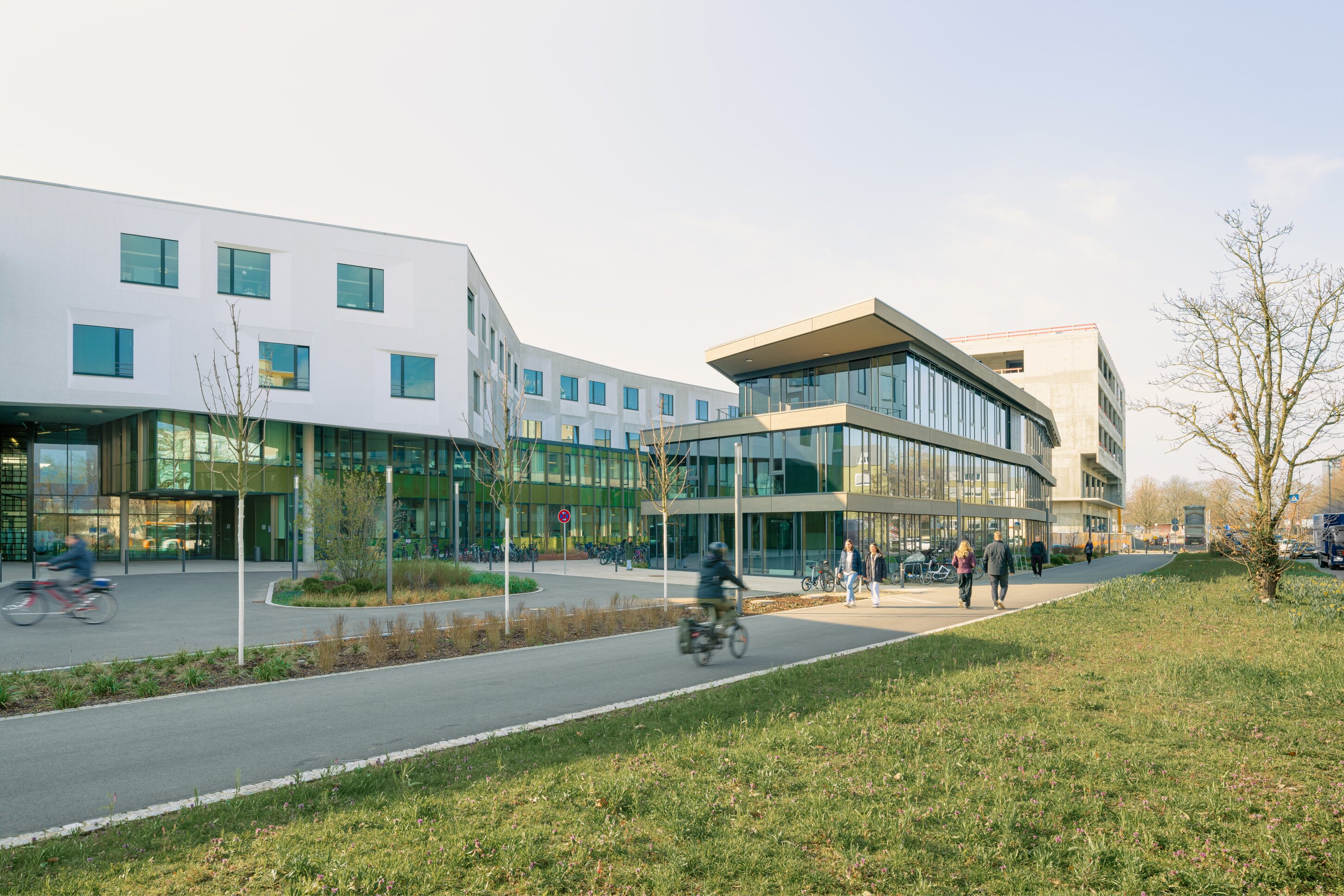 Project by Behnisch Architekturbuero, NCT New Building and Expansion. Modern campus buildings with glass facades, people walking and cycling on a path, and a grassy area with trees in the foreground.