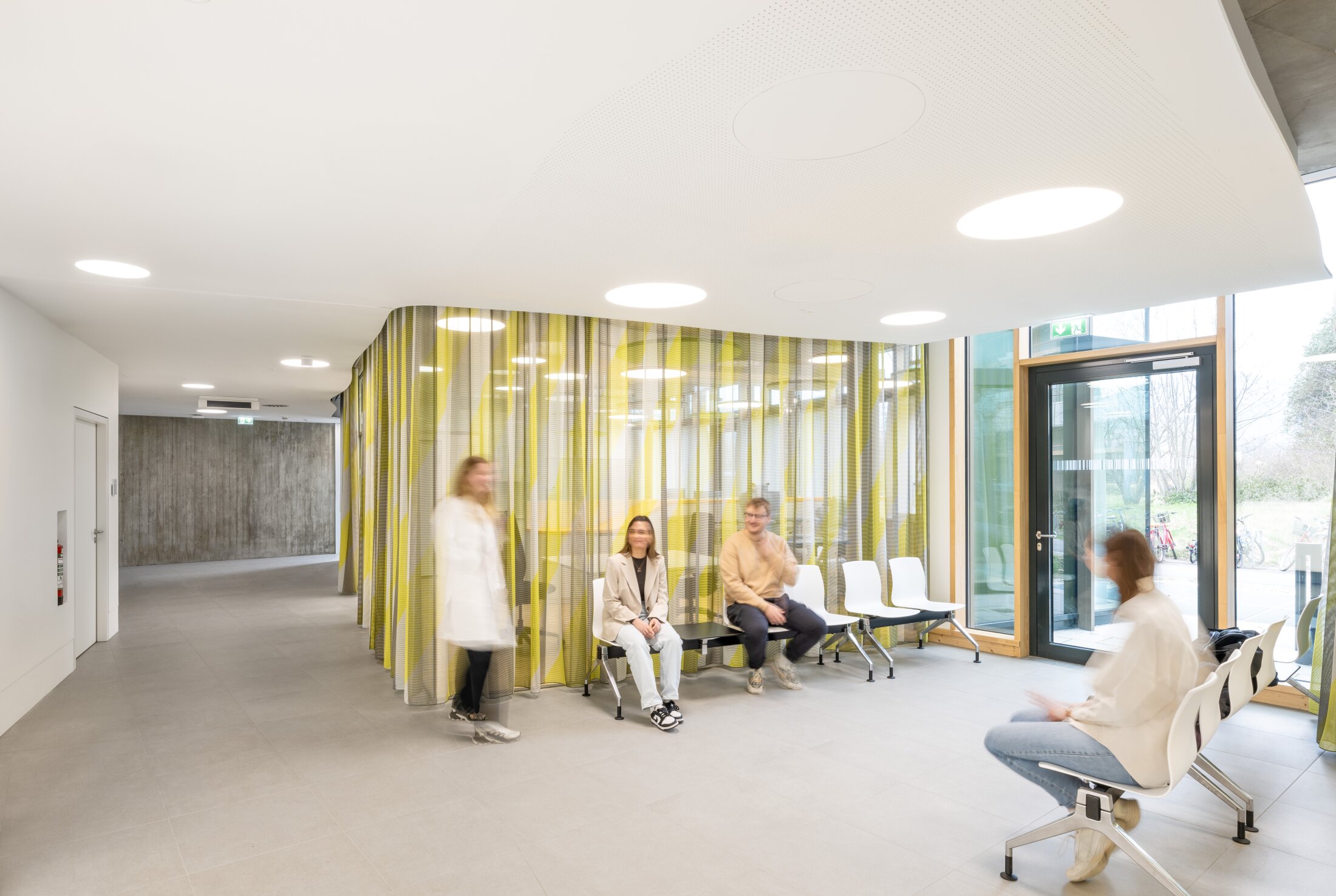 Project by Behnisch Architekturbuero, NCT New Building and Expansion. A bright and inviting waiting area with chairs, people sitting and walking around, and a transparent yellow curtain dividing a bright interior.