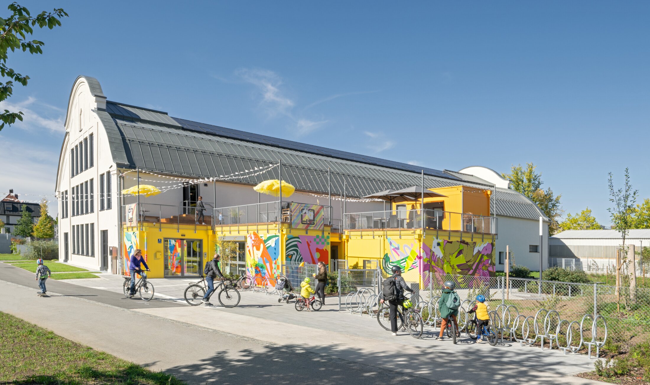 Project by Behnisch Architekturbuero, Action Sport Center. A hall with a container-like extension, a terrace, and people with bicycles in front of it. The containers are yellow and also painted in bright colors.