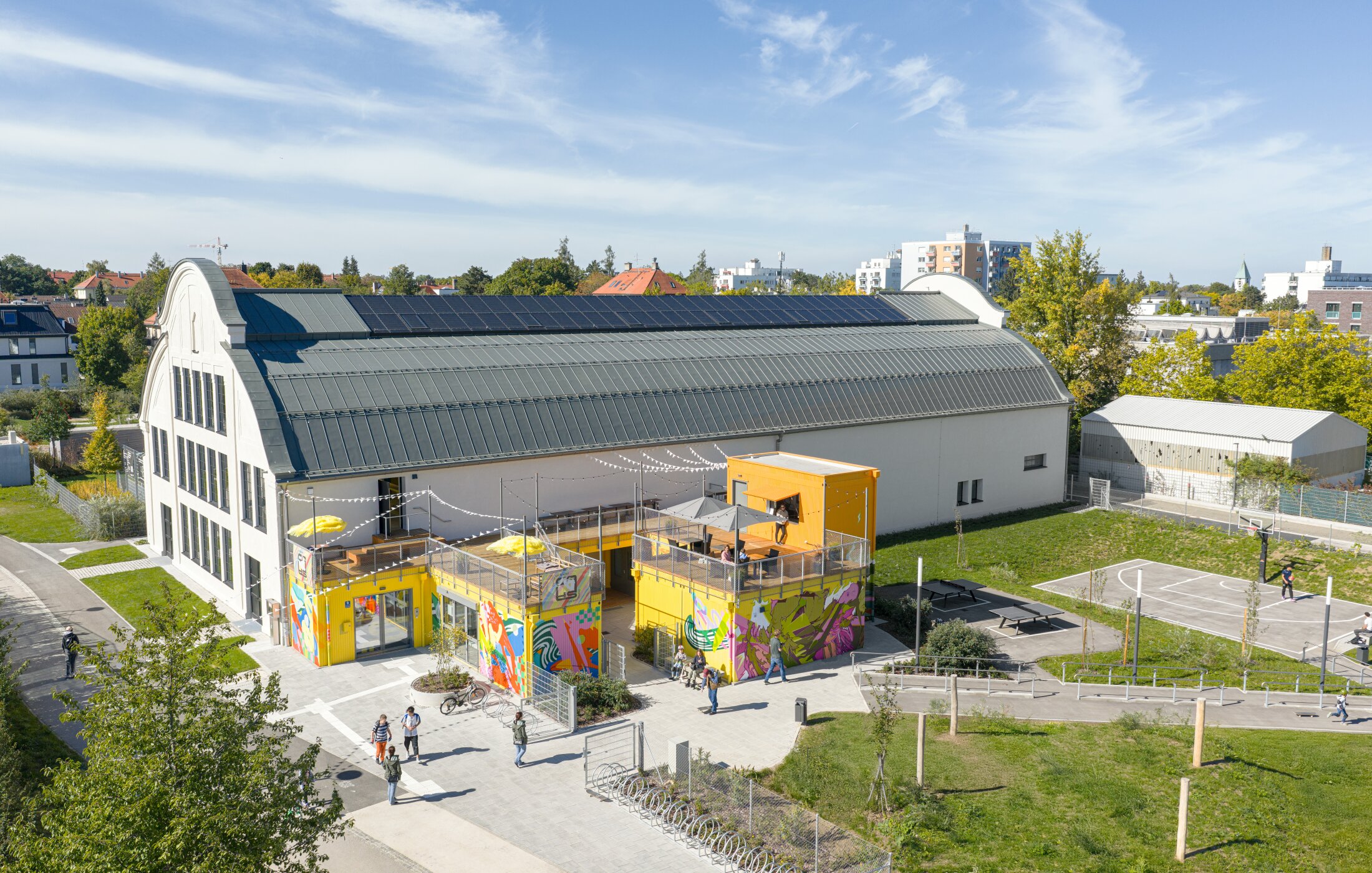 Project by Behnisch Architekturbuero, Action Sport Center. A hall with a container-like extension, a terrace, and people with bicycles in front of it. The containers are yellow and also painted in bright colors.