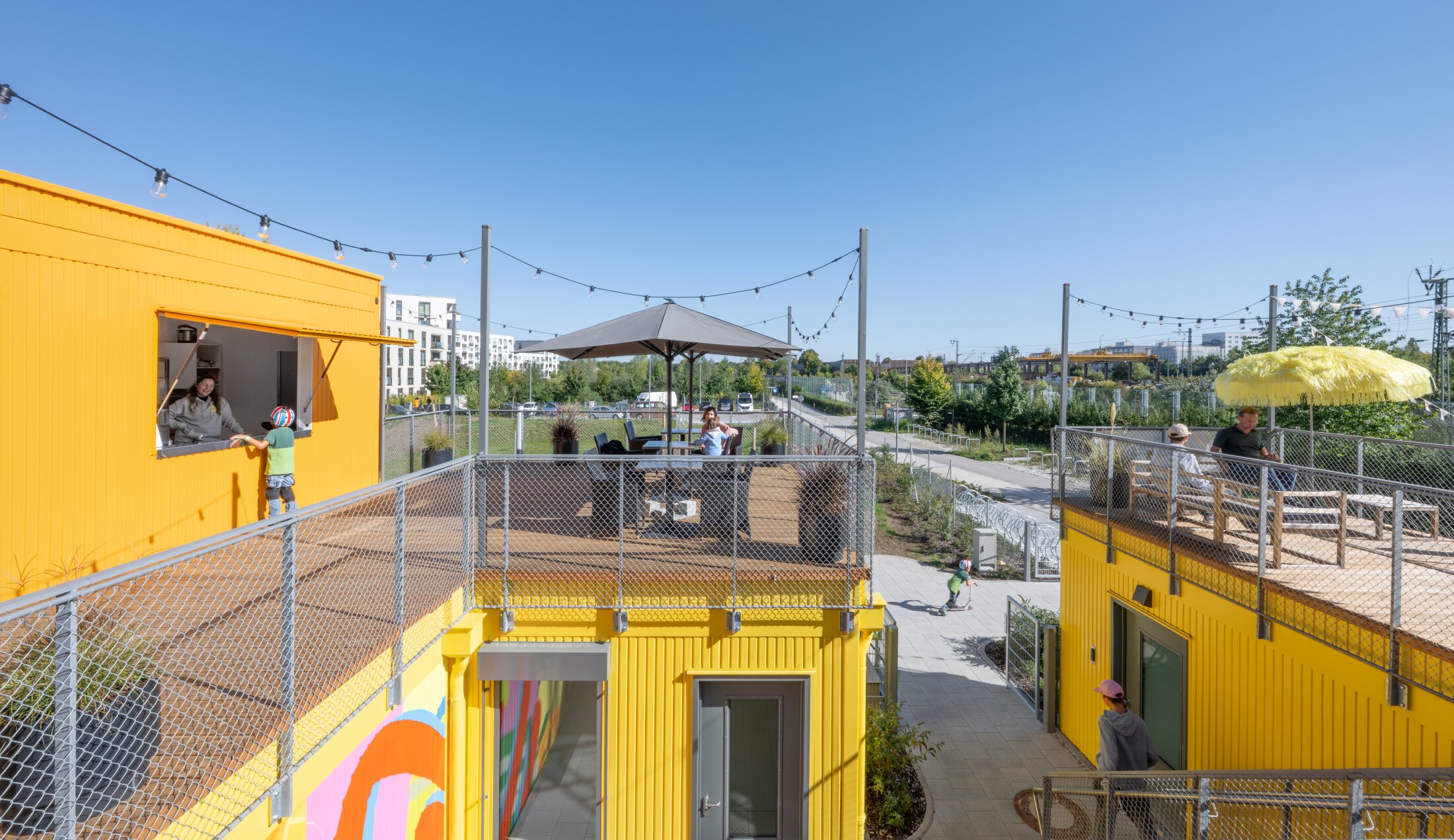 Project by Behnisch Architekturbuero, Action Sport Center. Yellow container-like terraced buildings with seating, people and strings of lights outdoors.