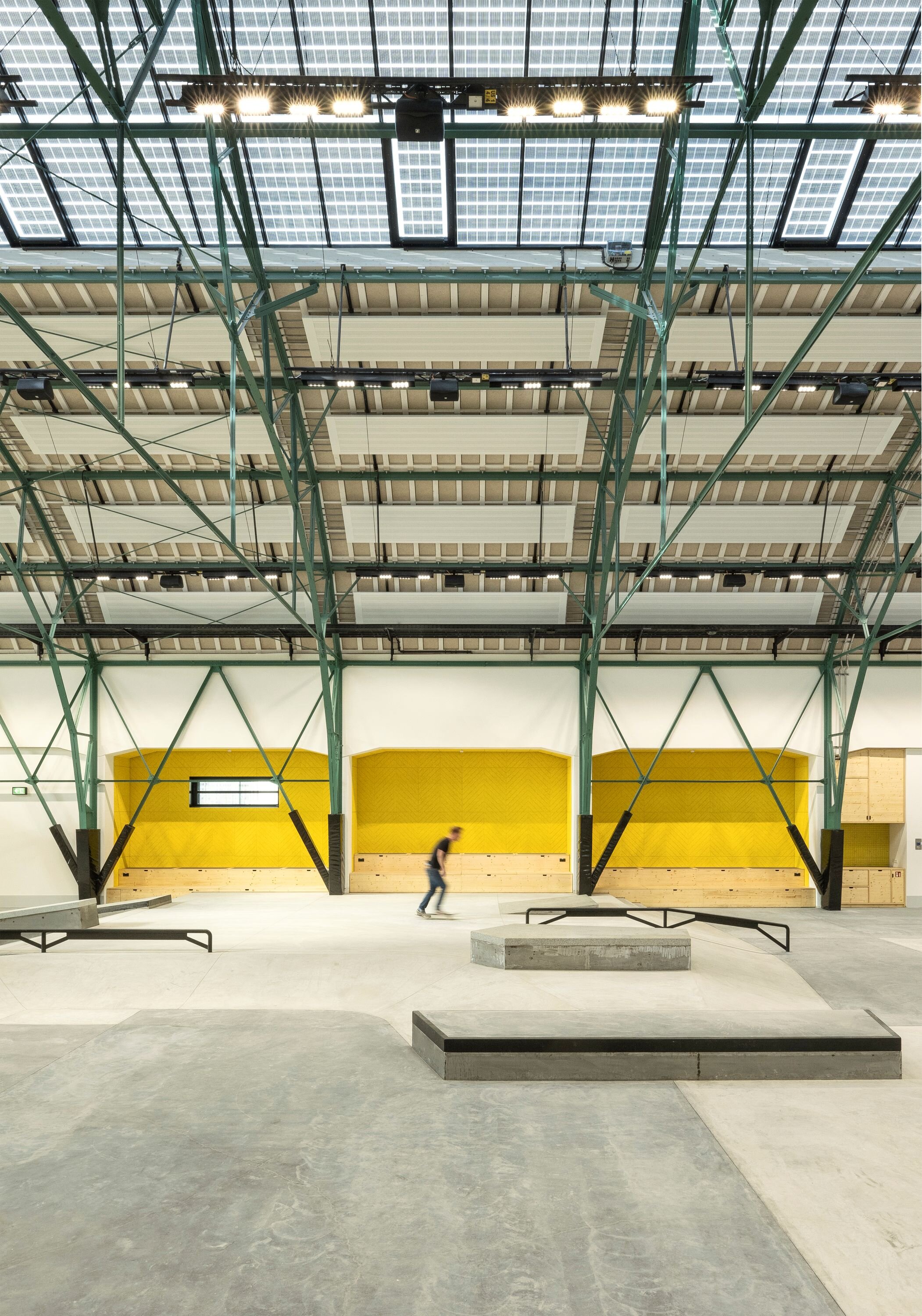 Project by Behnisch Architekturbuero, Action Sport Center. Indoor skatepark with ramps and rails under a steel truss roof, skaters in motion. Wooden seating and a yellow background.