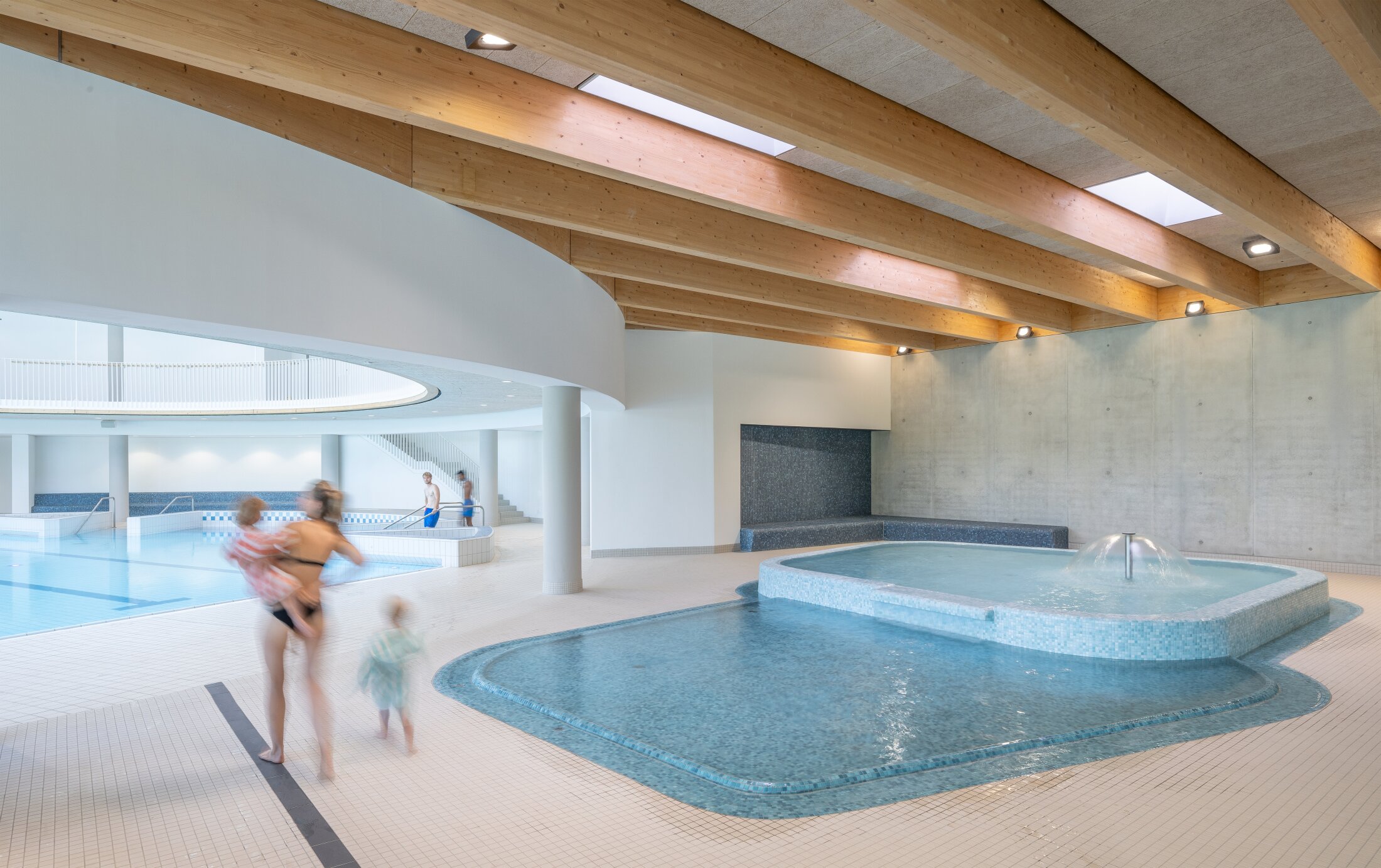 Project by Behnisch Architekturbuero, Wunnebad Winnenden. Indoor pool area with wooden beam ceiling, children and adults near shallow pool and fountain.