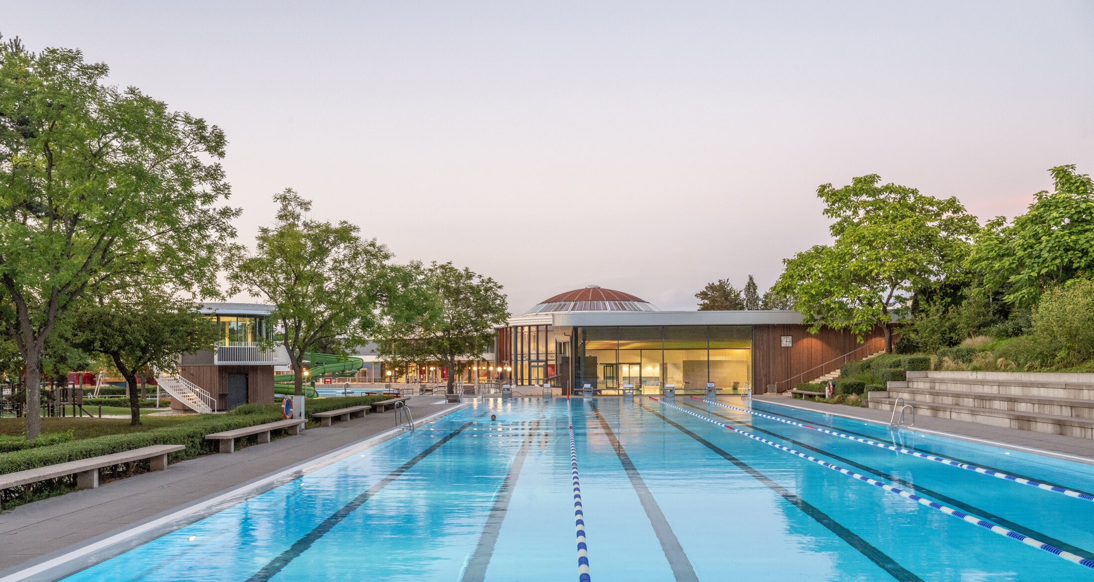 Project by Behnisch Architekturbuero, Wunnebad Winnenden. An outdoor swimming pool with lanes, trees, seating, and low buildings with domed roofs. On the right of the picture, steps can be seen, which also serve as seating.