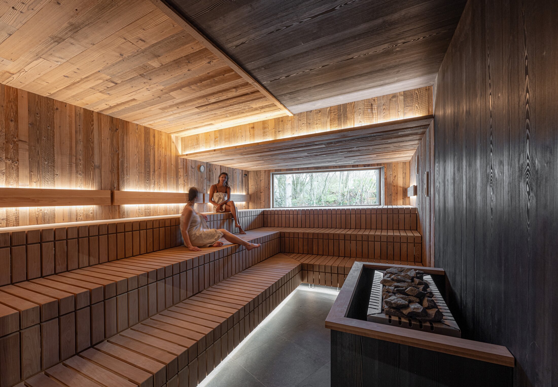 Project by Behnisch Architekturbuero, Wunnebad Winnenden. Two women are sitting in a wood-paneled sauna. The sauna stove can be seen on the right in the foreground. There is a window at the back of the sauna with a view outside.