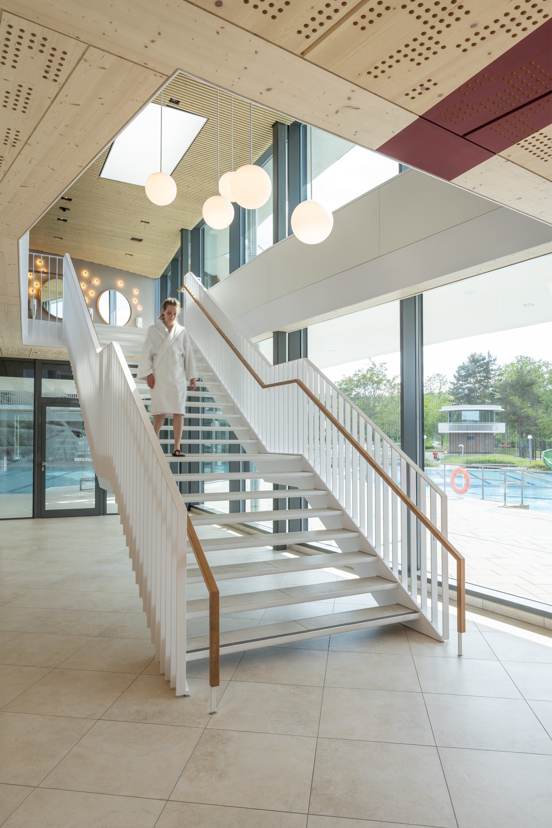 Project by Behnisch Architekturbuero, Wunnebad Winnenden. A person in a bathrobe descends the stairs to the swimming area. It is a bright, light-filled room with a wooden ceiling and large windows overlooking the outside.