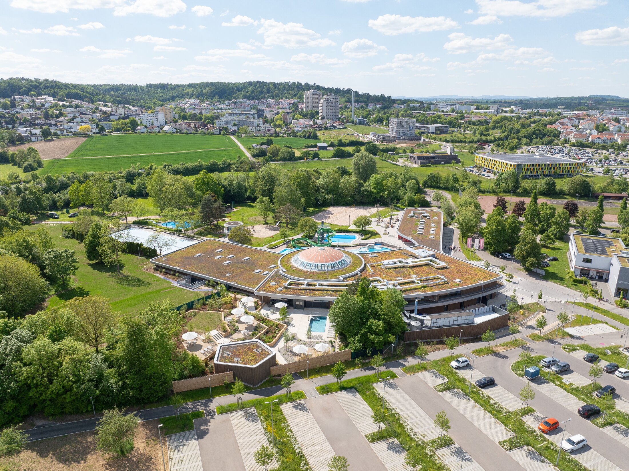 Project by Behnisch Architekturbuero, Wunnebad Winnenden. Aerial view of the building and its exterior. The outdoor spa and swimming pool area are visible. The spa's parking lot can be seen in the lower part of the image.