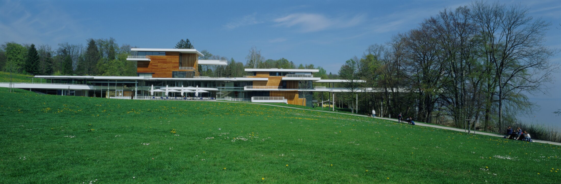Project by Behnisch Architekturbuero, Buchheim Museum. A modern building with wooden facades on a green hillside, with trees and people nearby. A lake can be seen in the background on the right.