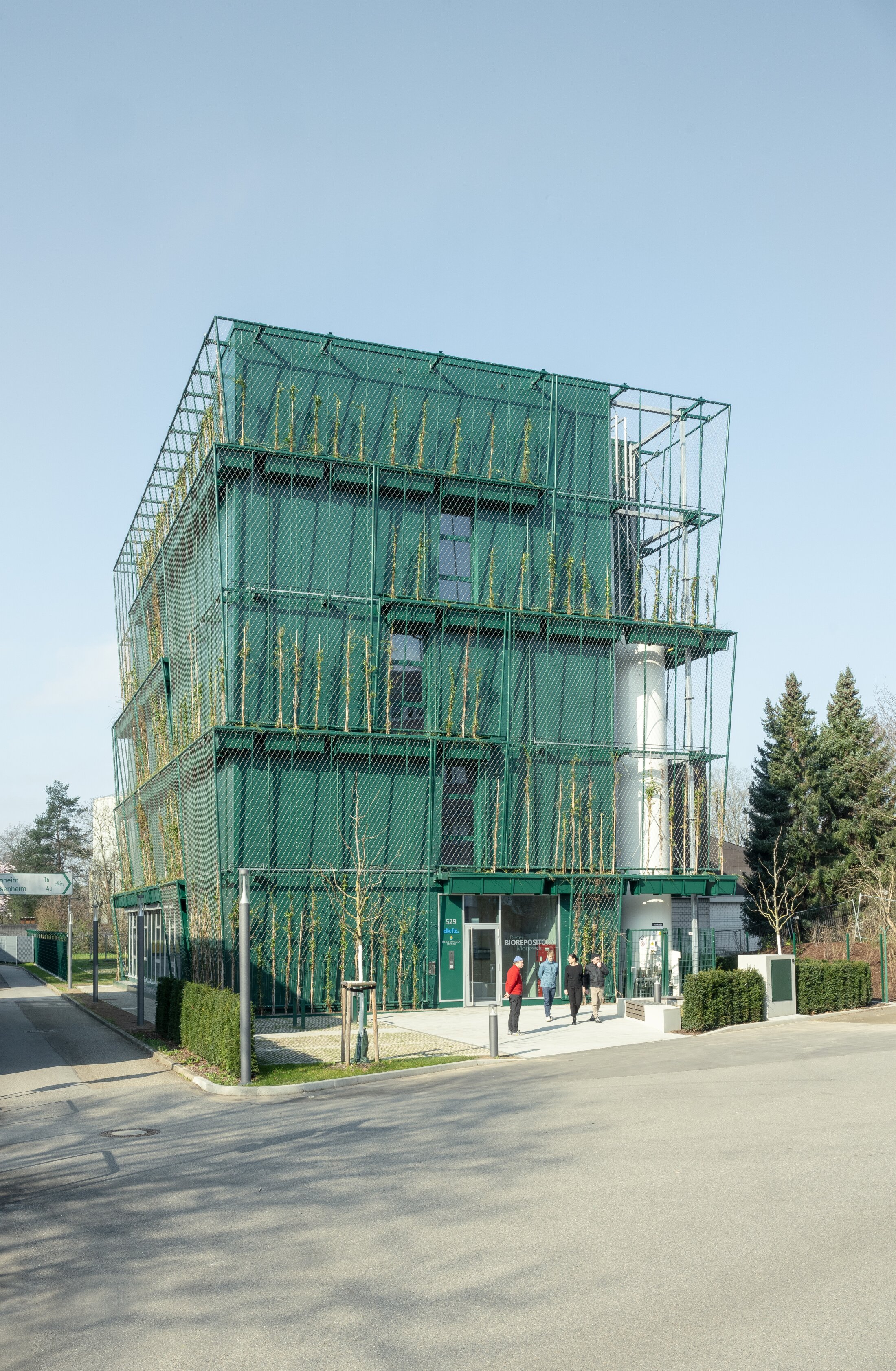 Project by Behnisch Architekturbuero, Dieter Morszeck Biorepository. Corner view of green multi-story building with green facade and climbing plants, people near entrance.