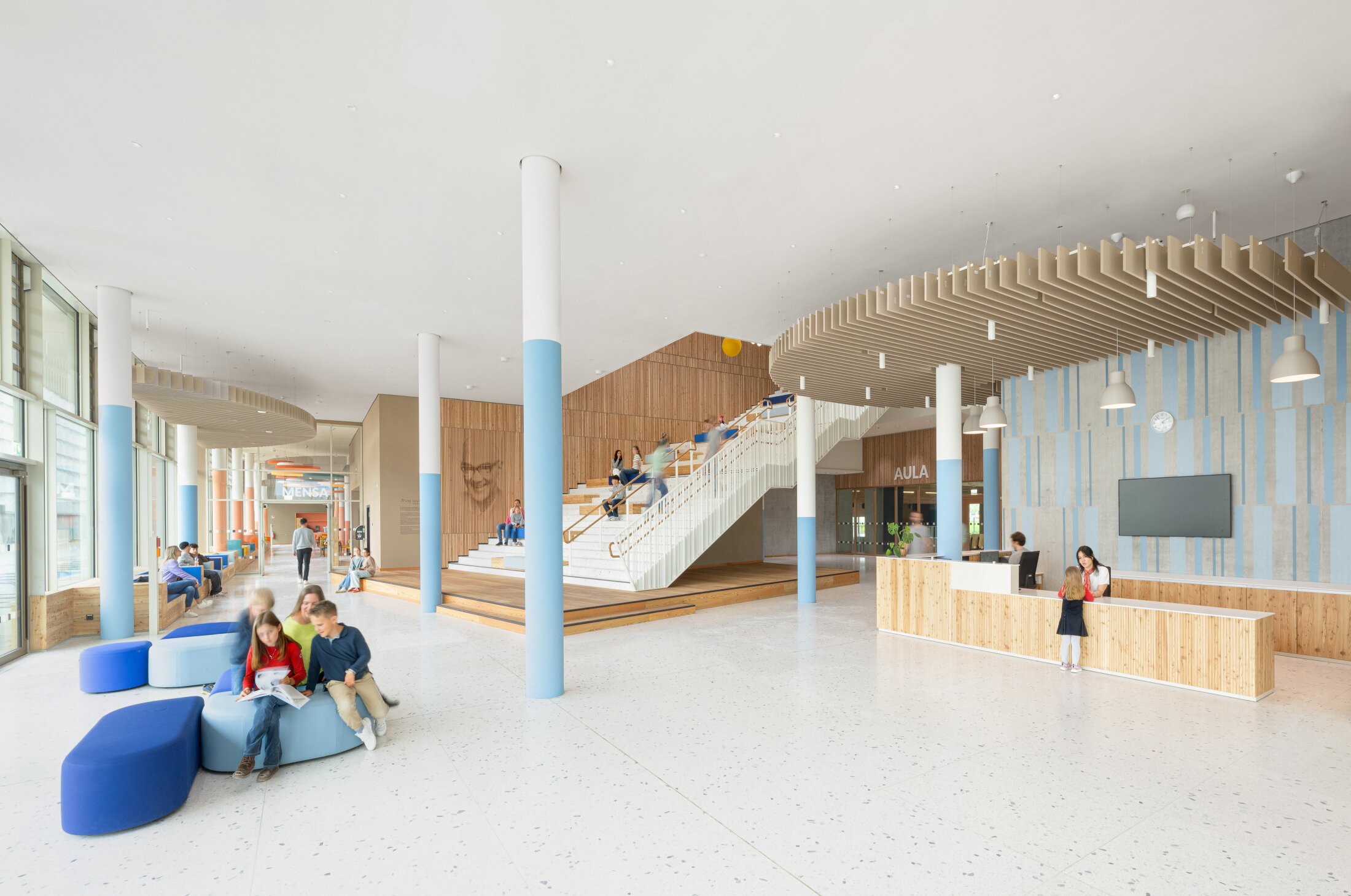 Project by Behnisch Architekturbuero, Josef-Schwarz School. The students gather in a bright school hall with stairs, seating, a reception area, and large windows. Children sit on the dark and light blue chairs and read.