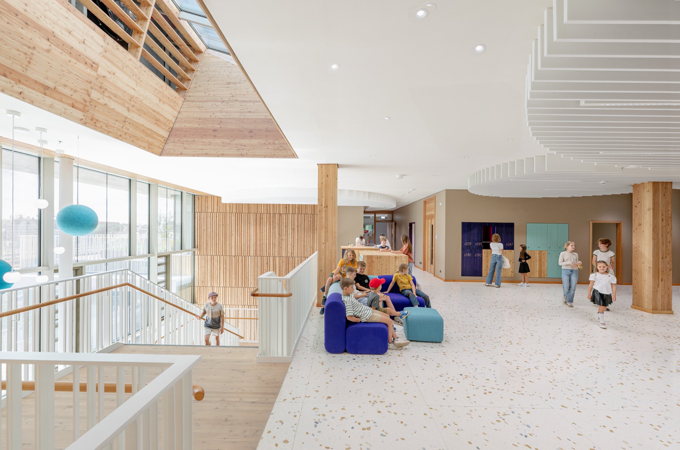 Project by Behnisch Architekturbuero, Josef-Schwarz School. Children and adults in a bright school lobby with stairs, seating, and wooden finishes.
