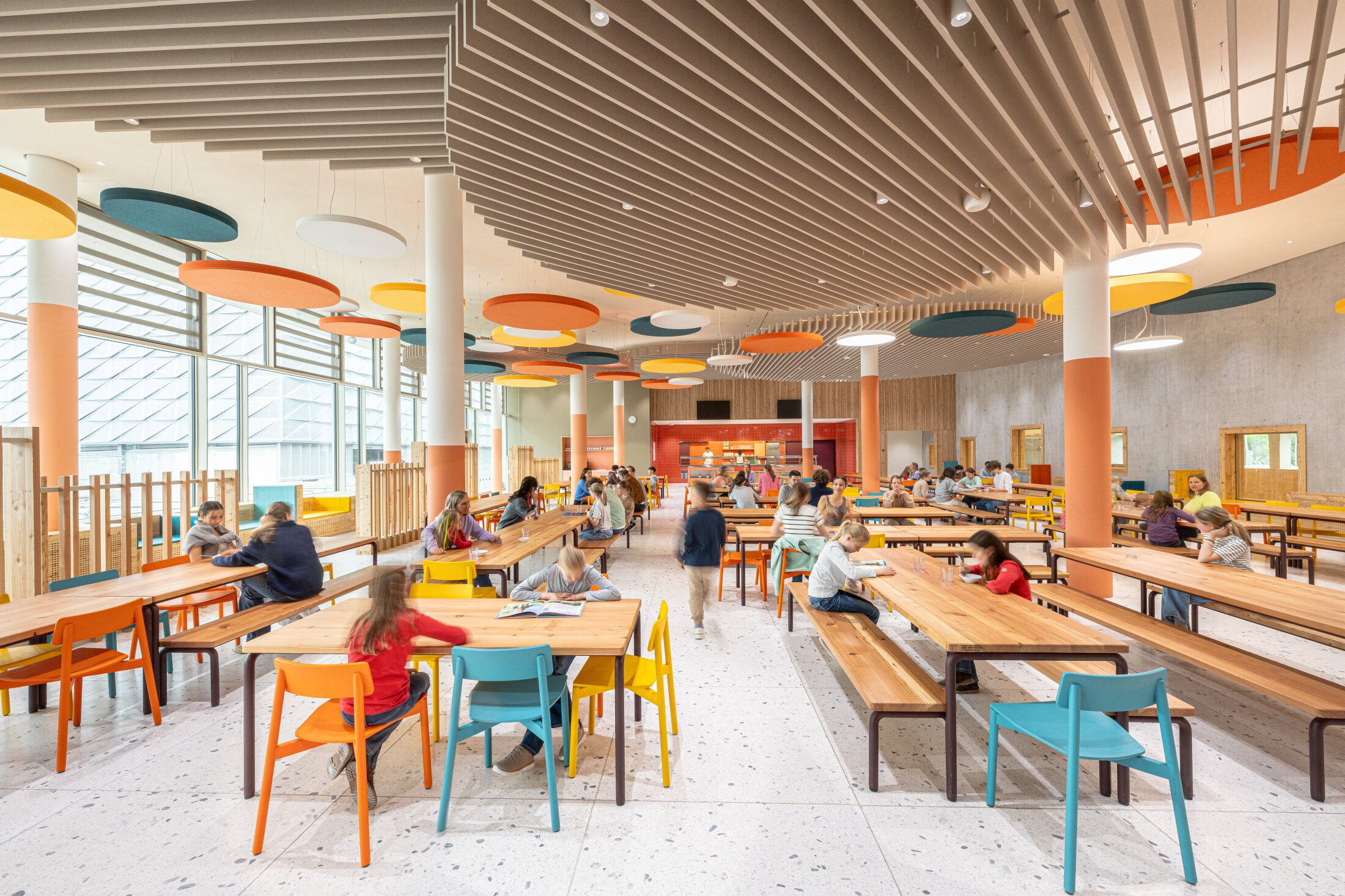 Project by Behnisch Architekturbuero, Josef-Schwarz School. The students sit at long tables in a bright cafeteria with colorful chairs, large windows and colorful acoustic ceiling panels.