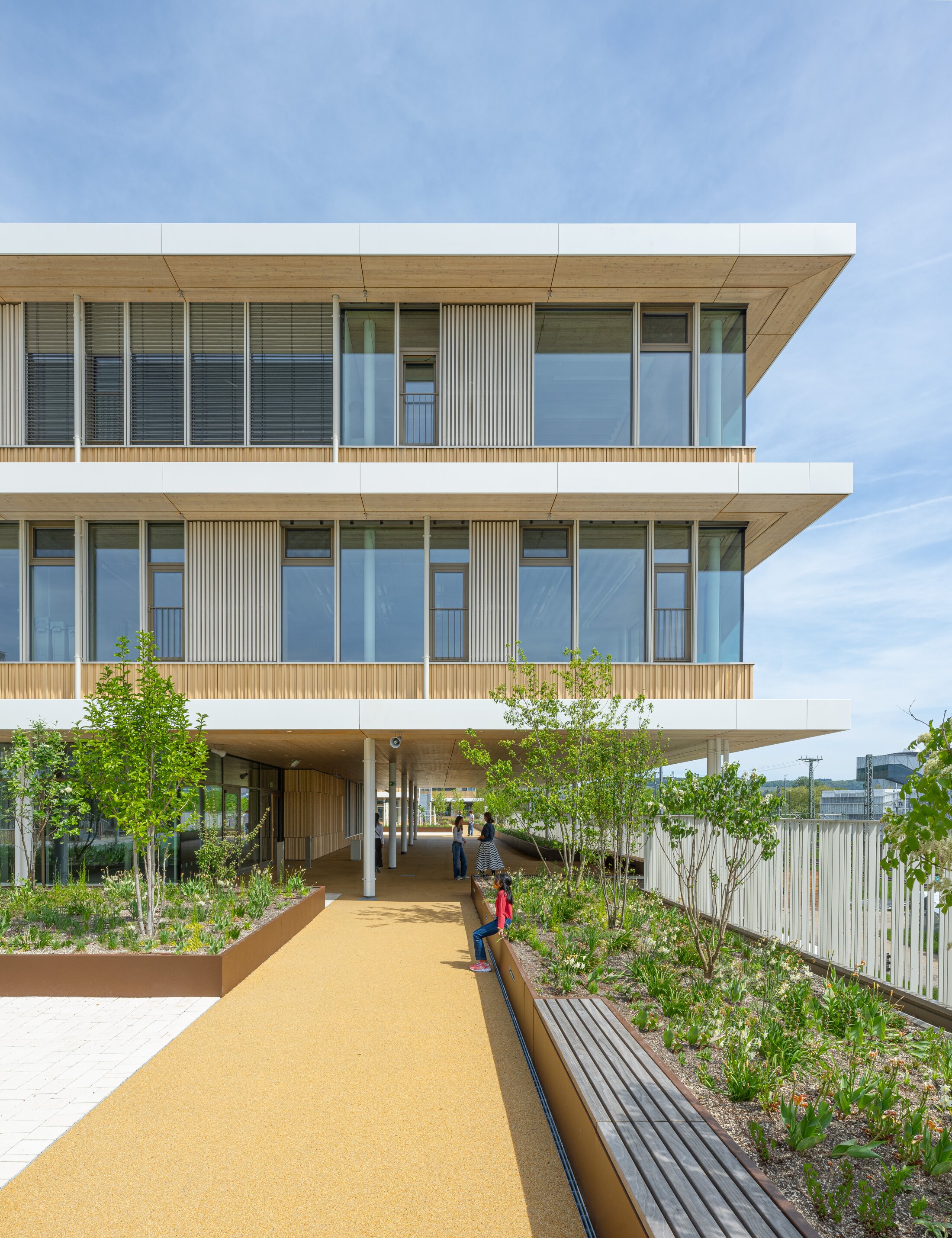 Project by Behnisch Architekturbuero, Josef-Schwarz School. Modern wood-clad building with large windows, shaded walkway, and landscaped path with trees and seating.