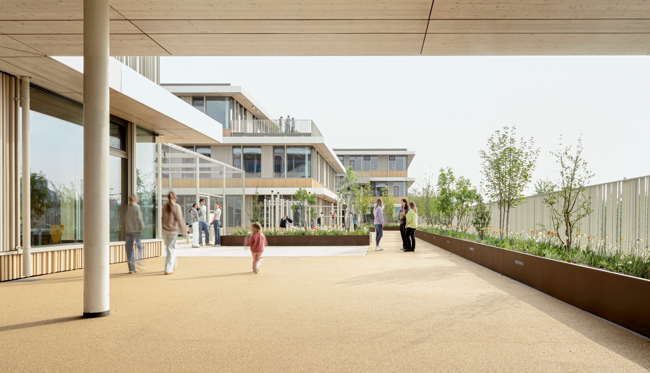 Project by Behnisch Architekturbuero, Josef-Schwarz School. Students and teachers are in a green outdoor area between the modern buildings. A raised bed with plants and trees can be seen on the right. A climbing frame is visible in the background on the left.