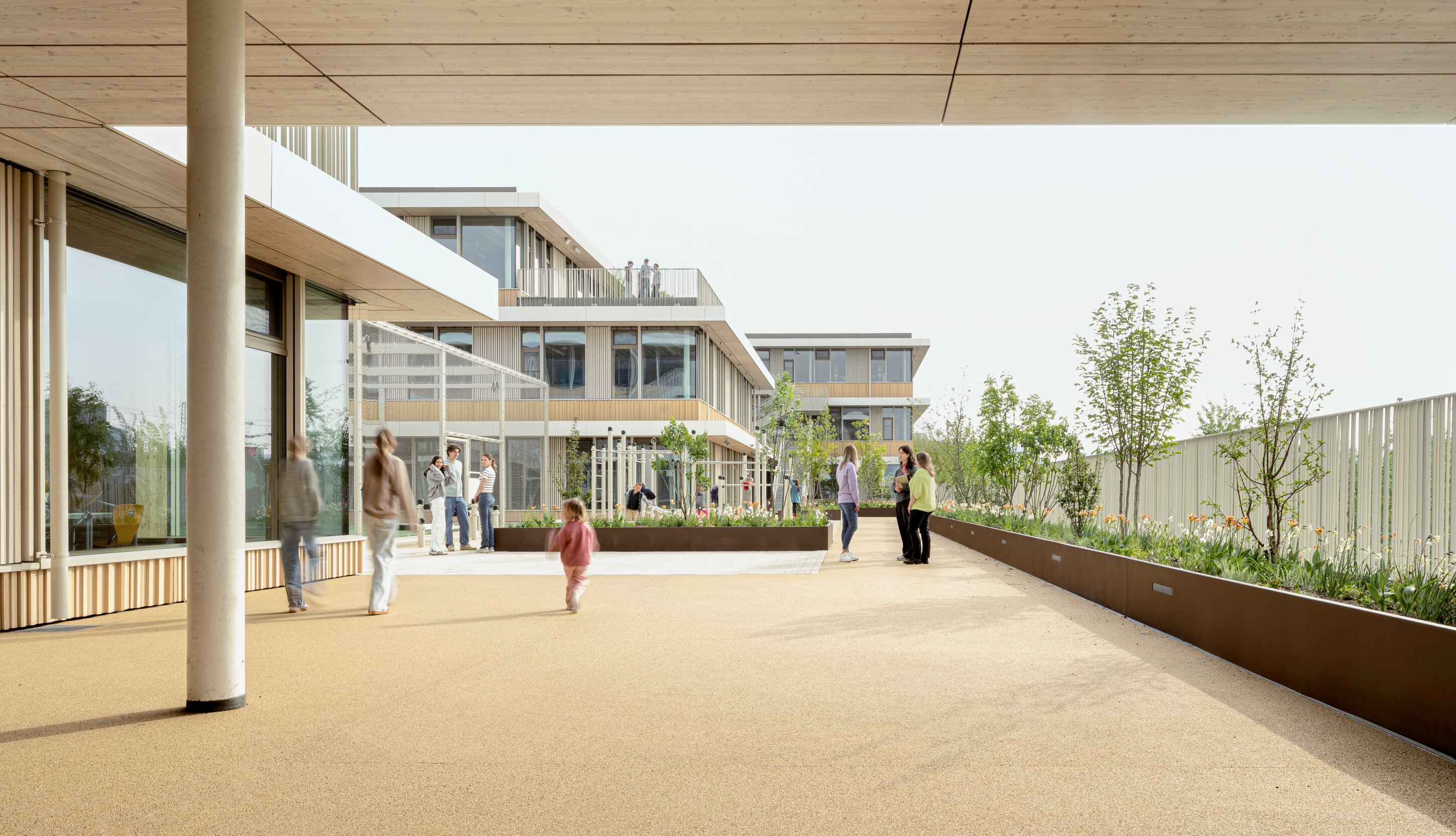 Project by Behnisch Architekturbuero, Josef-Schwarz School. Students and teachers are in a green outdoor area between the modern buildings. A raised bed with plants and trees can be seen on the right. A climbing frame is visible in the background on the left.