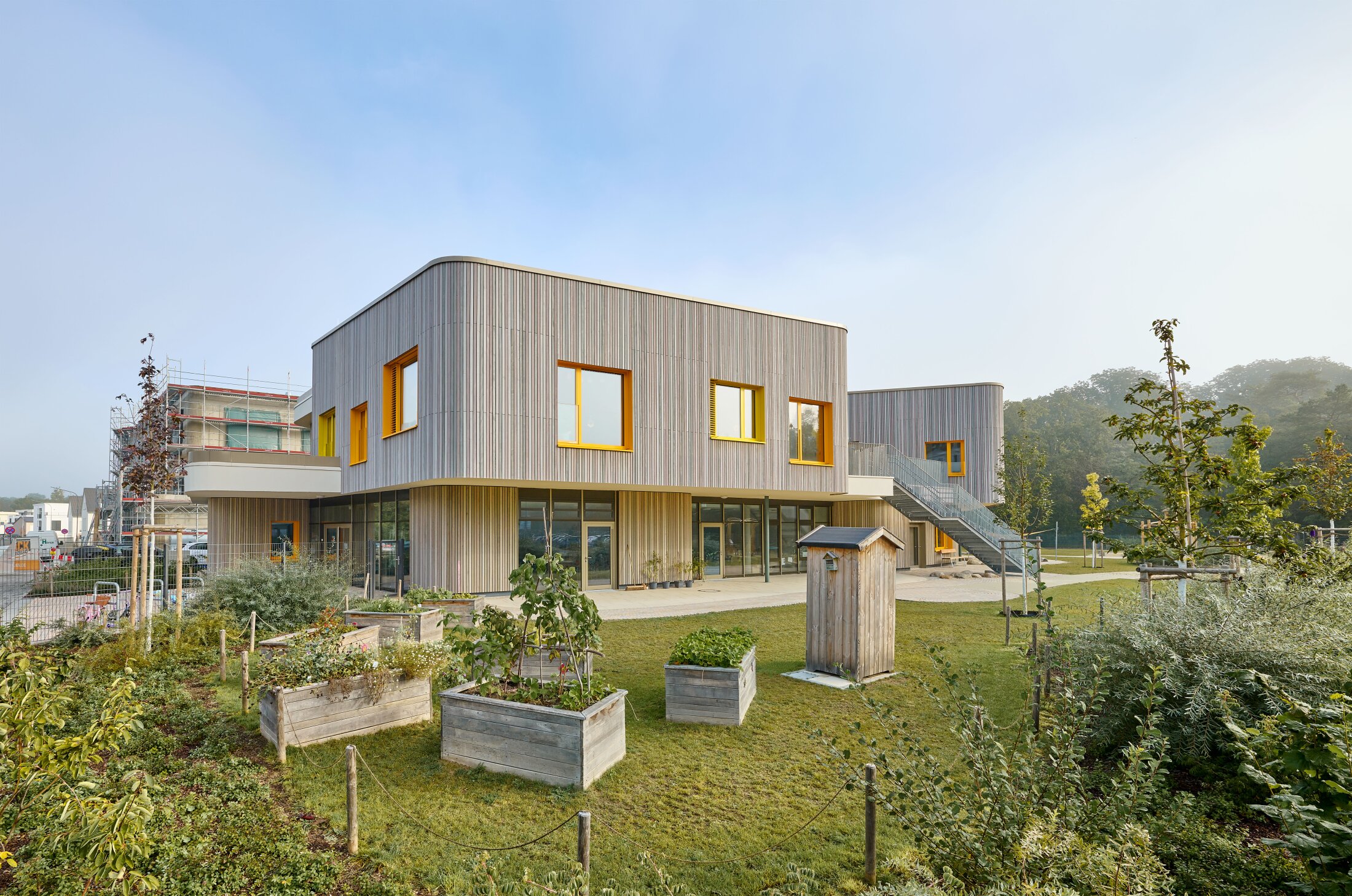 Project by Behnisch Architekturbuero, KiTa Pioneer-Kaserne. Modern wooden building with yellow and orange framed windows. Surrounded by a garden with raised beds and lawns.