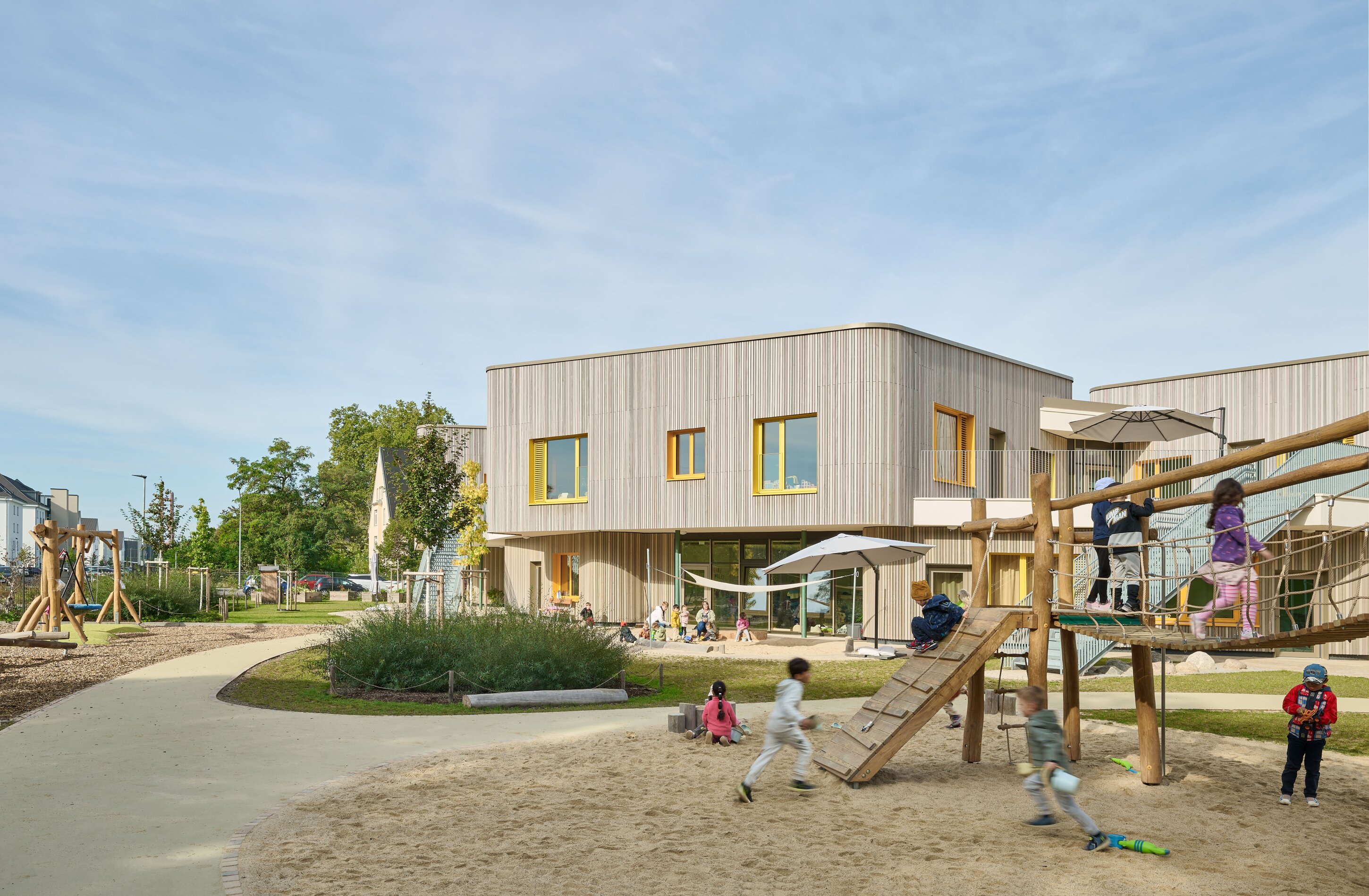 Project by Behnisch Architekturbuero, KiTa Pioneer-Kaserne. Children playing on a wooden playground structure in a sandy area beside a modern school building with a courtyard and greenery.