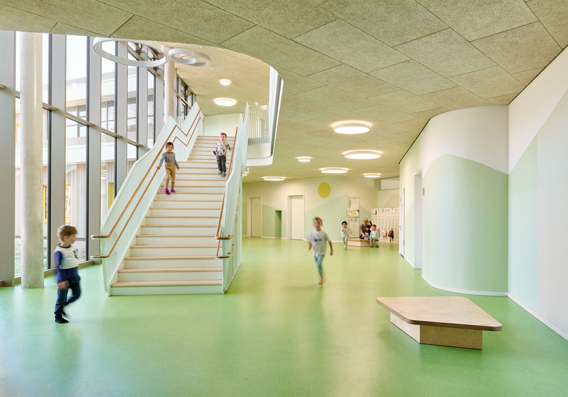Project by Behnisch Architekturbuero, KiTa Pioneer-Kaserne. Children play in a bright school interior with green floor, white staircase, and curved walls with windows and ceiling lights.